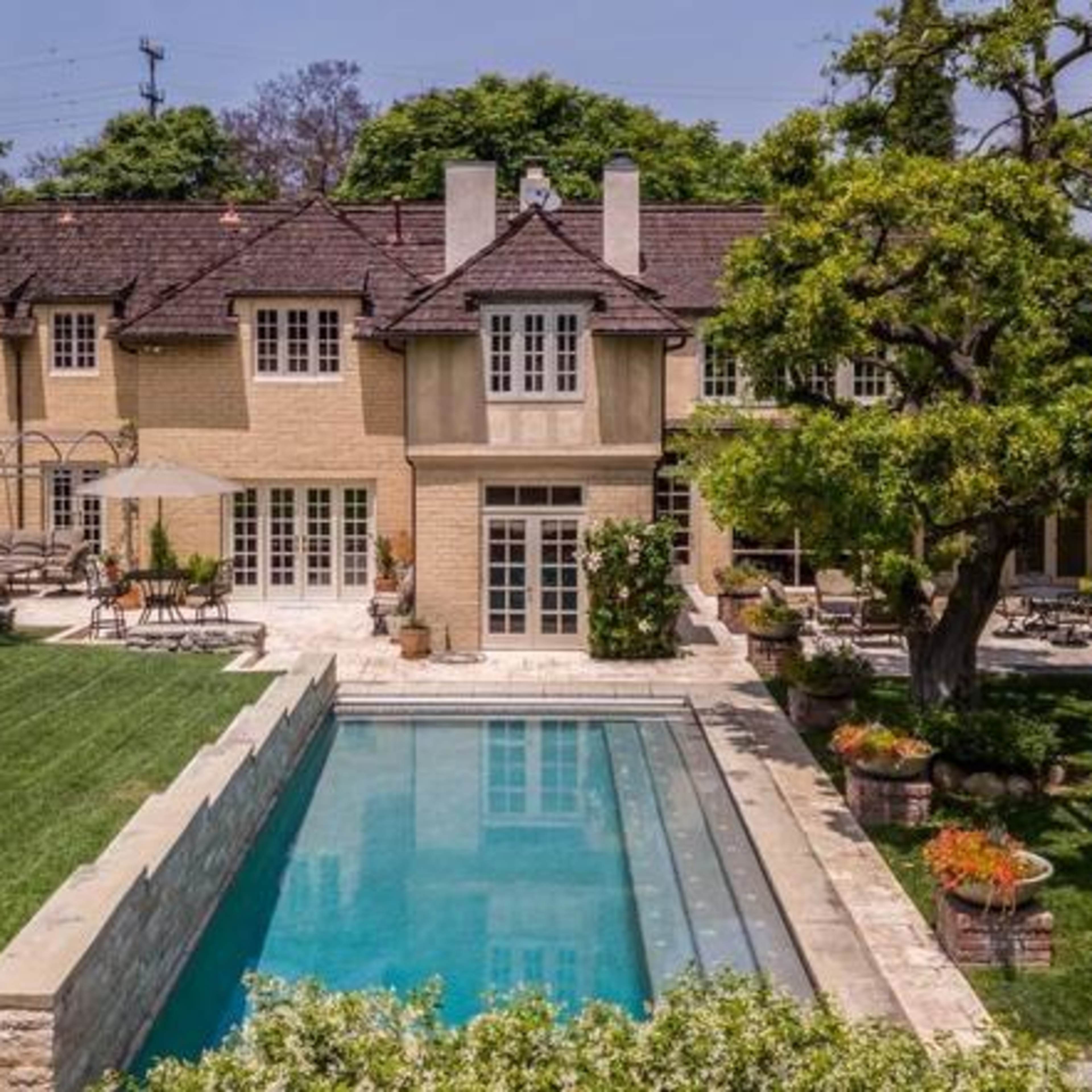 A large, multi-story house with a pool and landscaped lawn, surrounded by trees and lounge areas.