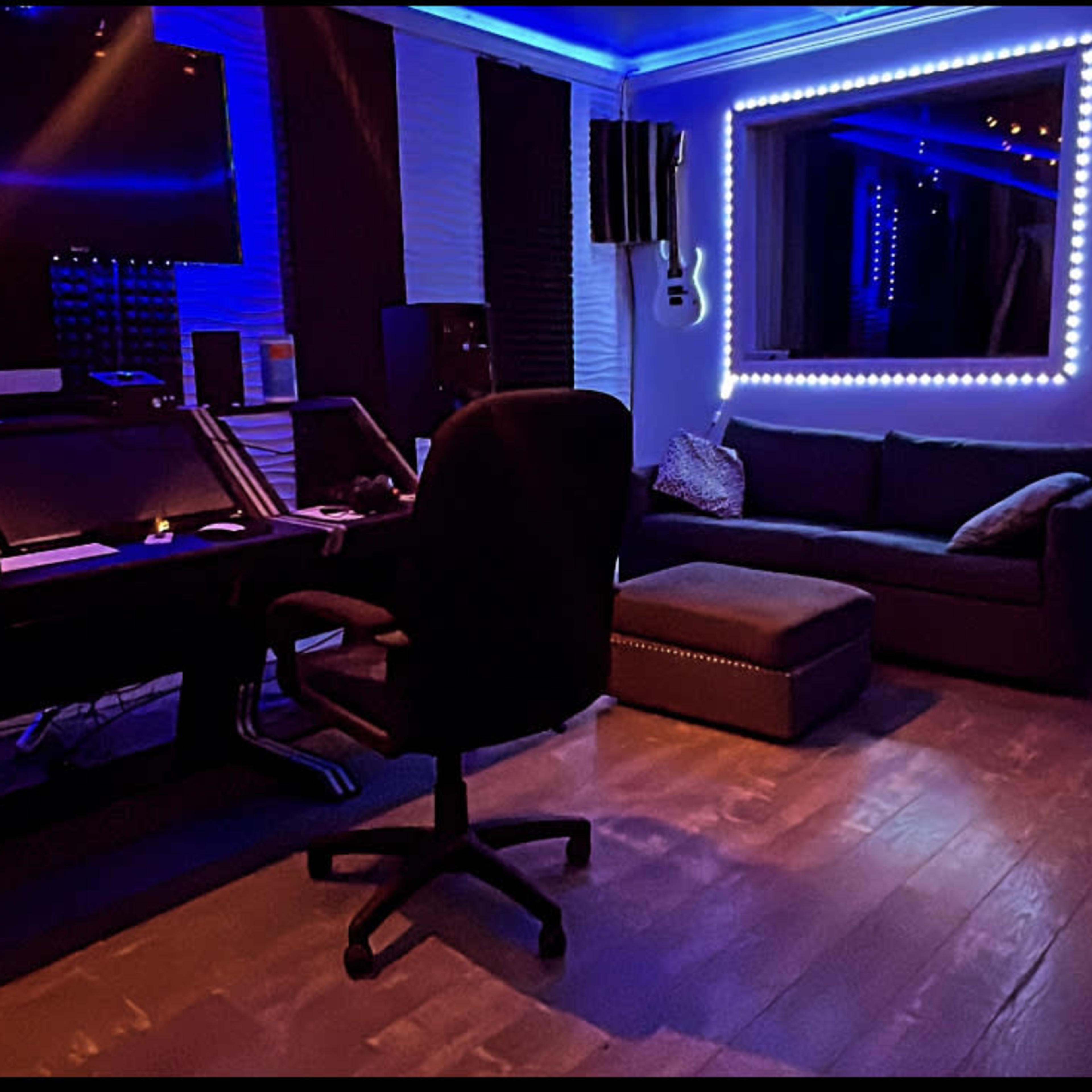 A music studio featuring a desk with computer equipment, a black swivel chair, a couch, and illuminated wall decorations.