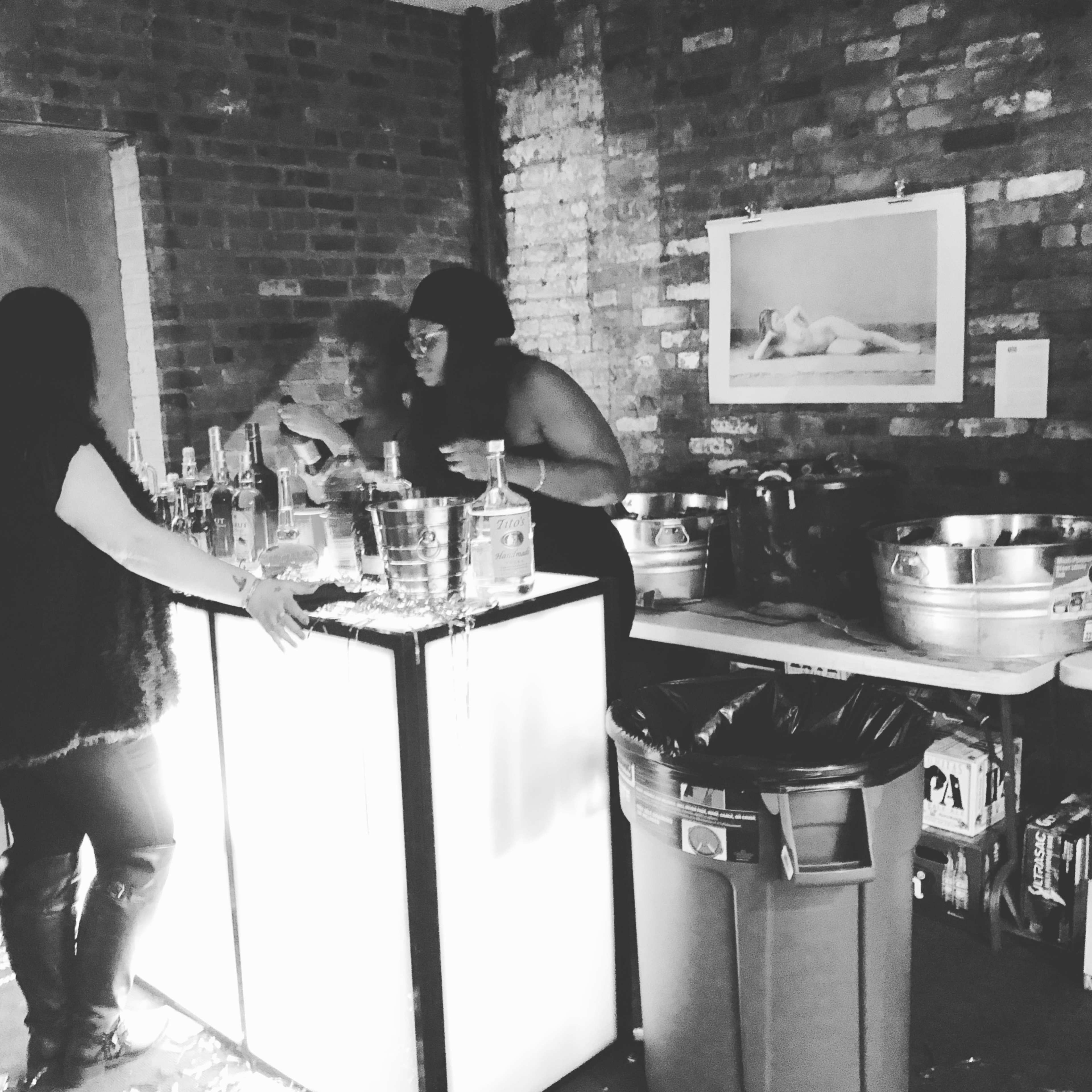 Two individuals stand behind a well-lit bar counter, surrounded by various bottles and bins, in a brick-walled room.
