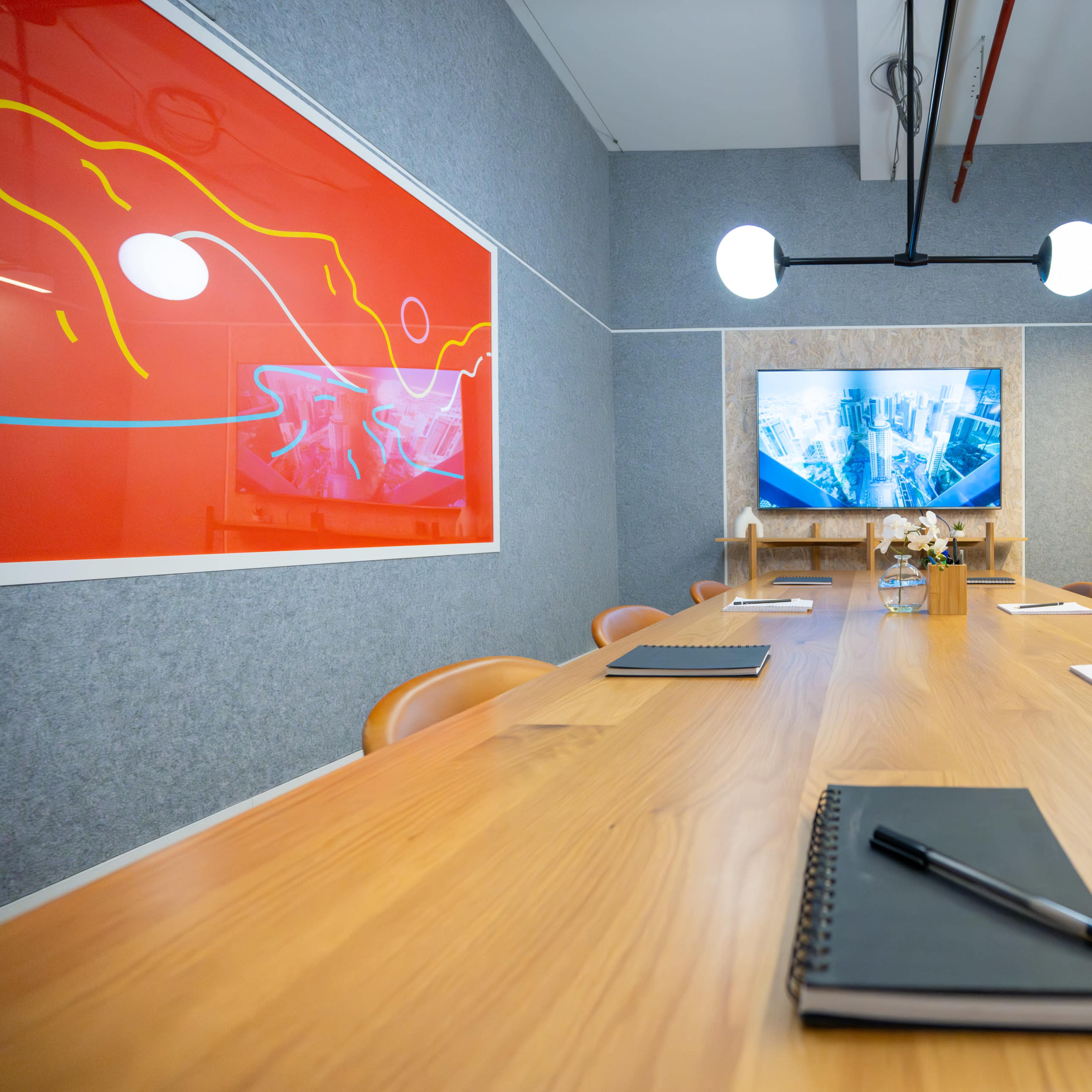 A modern conference room features a long wooden table with notebooks and pens, a large screen displaying a presentation, and colorful artwork on the walls.