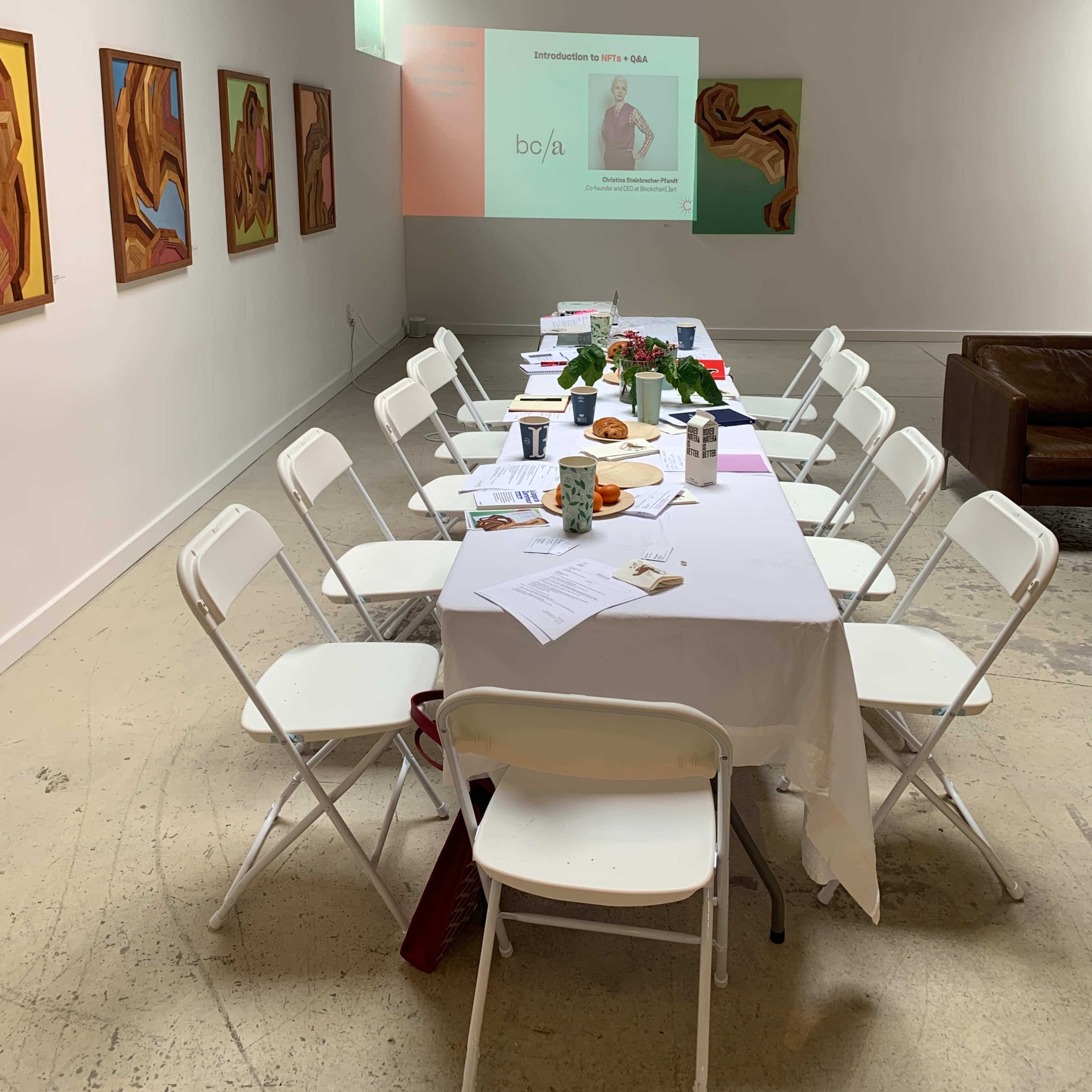 A long table with white chairs is set up in a bright room, featuring a presentation screen and several artworks on the walls.