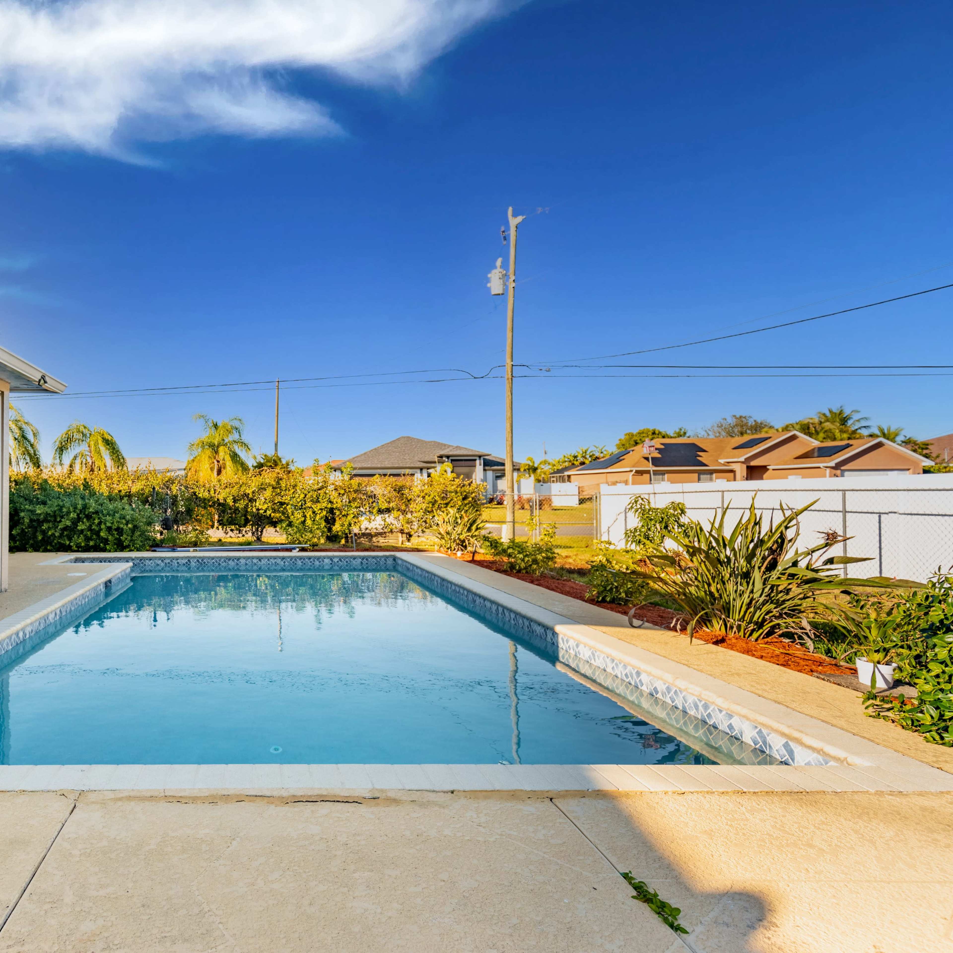 A clear swimming pool sits in an outdoor area surrounded by greenery and a fence under a bright blue sky.