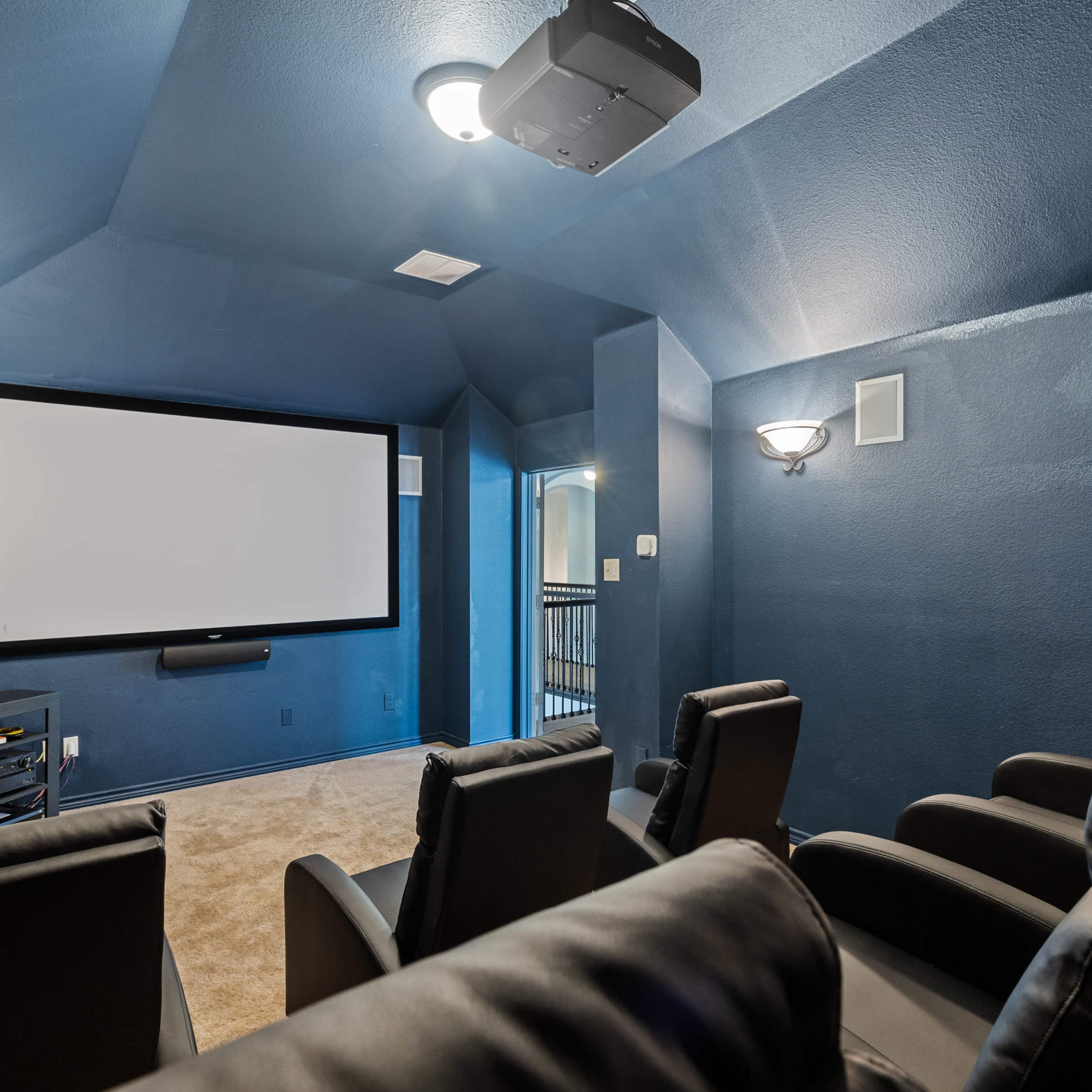 A dimly lit home theater features a large screen, a projector on the ceiling, and several black leather recliners arranged in front.