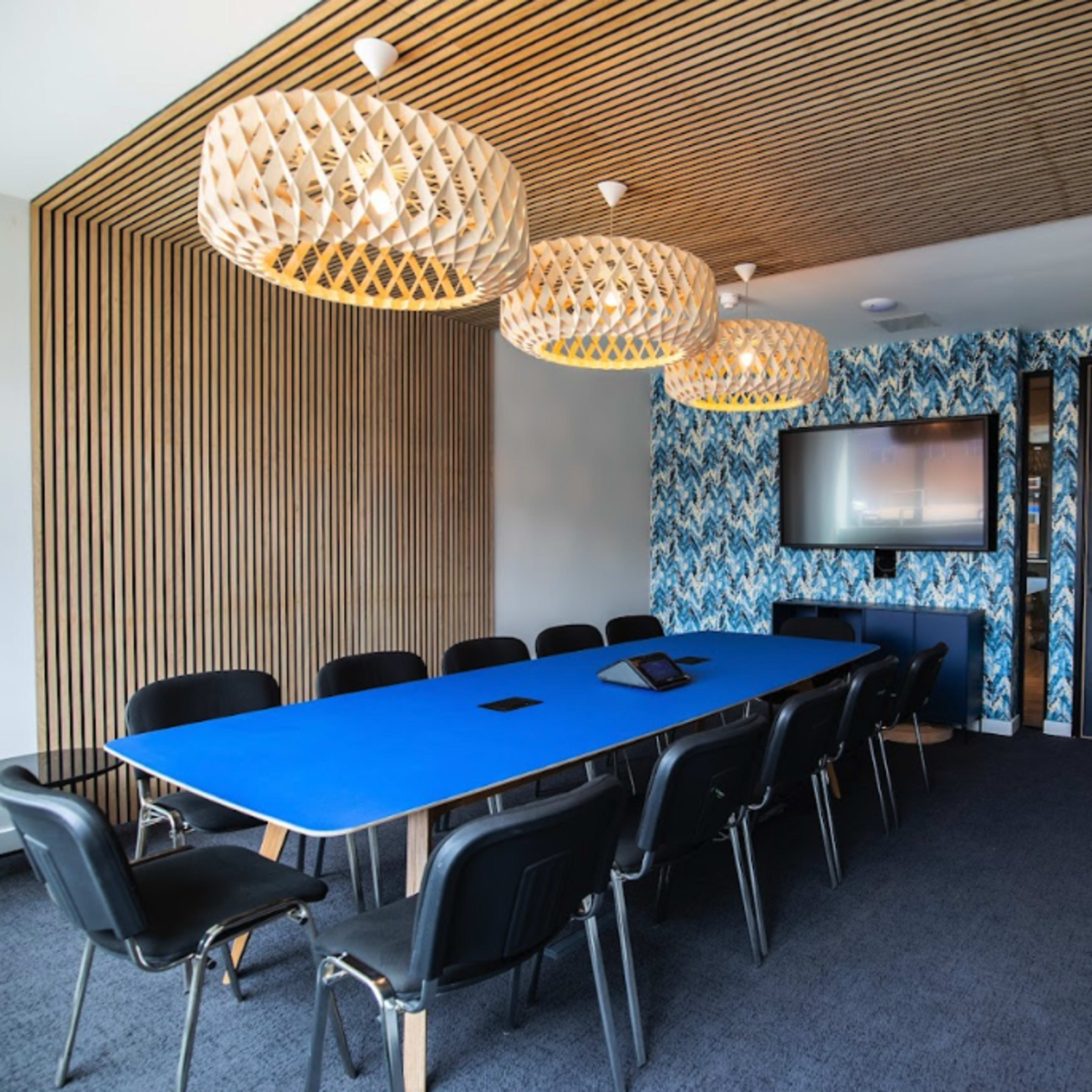 A modern conference room features a long blue table surrounded by black chairs, with patterned wallpaper and pendant lights above.