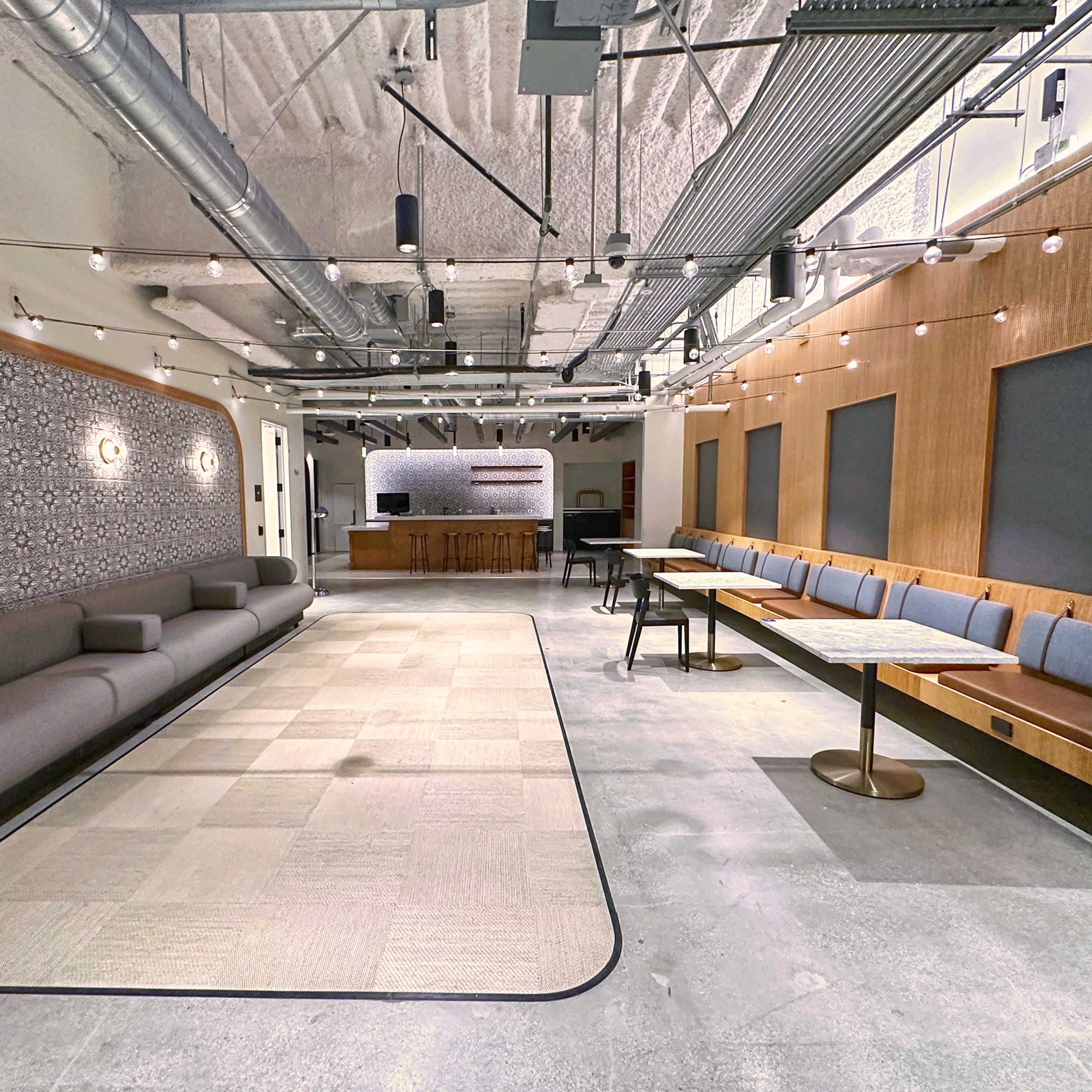 A spacious interior featuring a long gray sofa, a series of tables with wooden benches, and decorative lighting overhead.