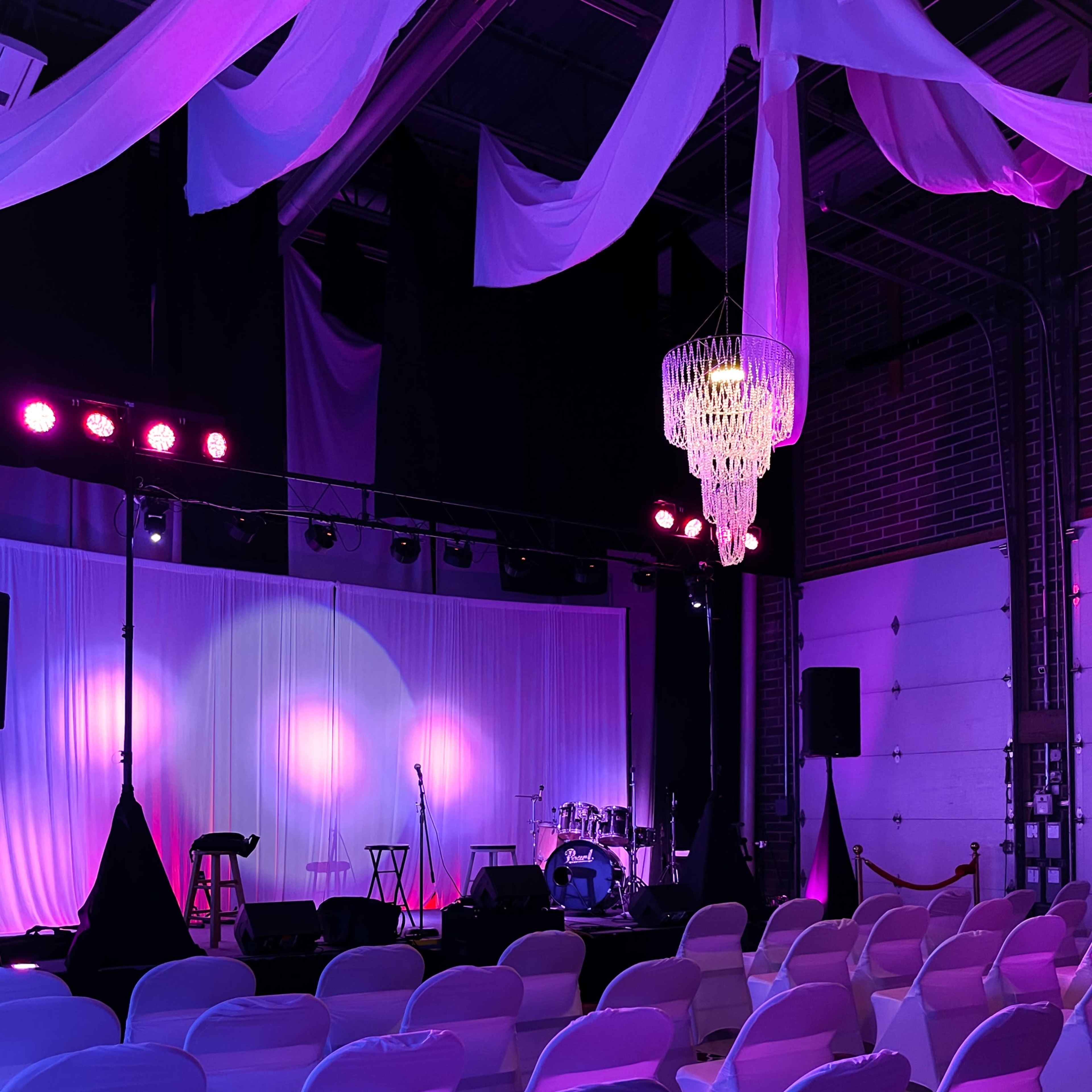 A stage is set up in a dimly lit venue, featuring a drum set and microphones, with rows of white chairs arranged for an audience.