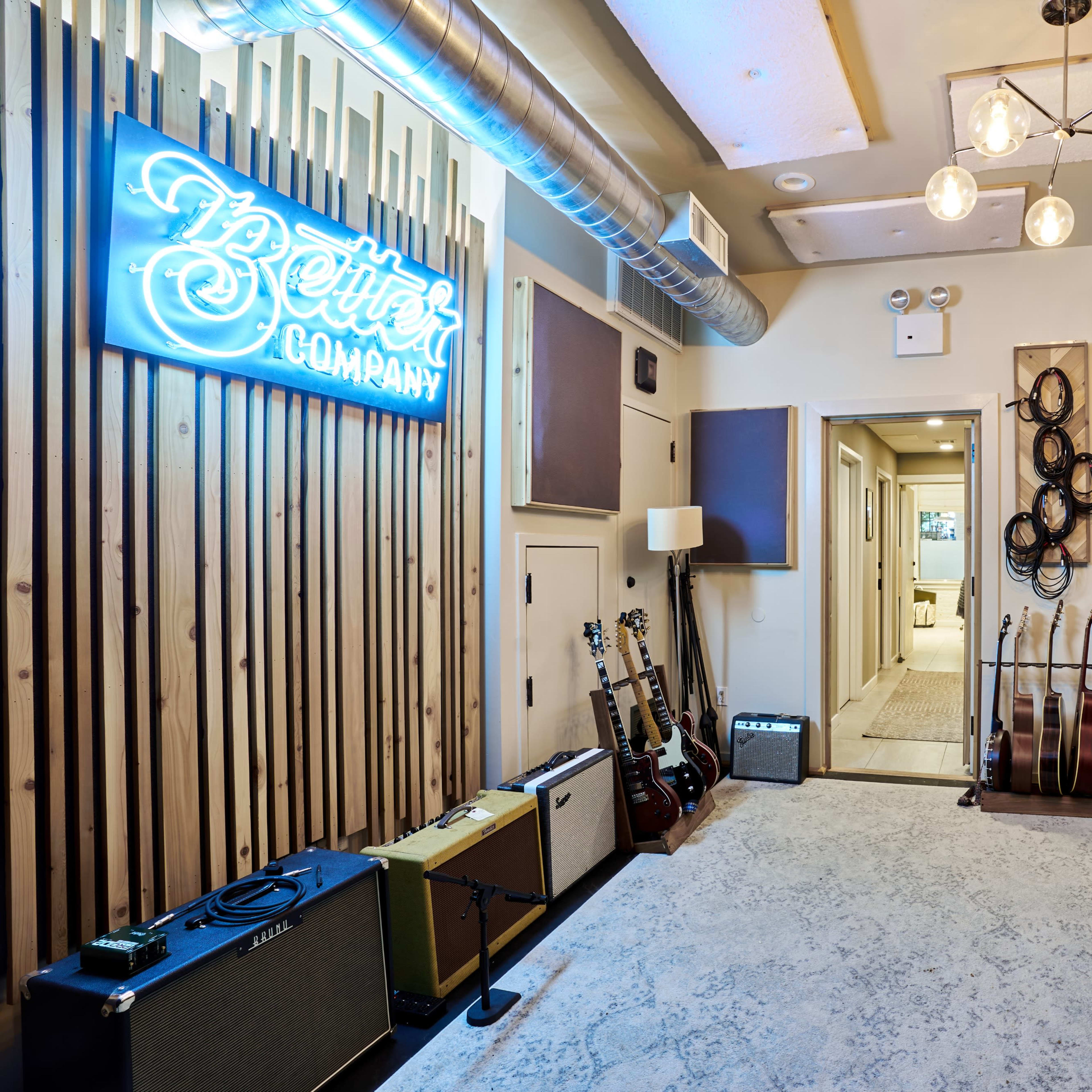 A music studio with wooden paneling, guitars, amplifiers, a piano, and a neon sign that reads "Better Company."