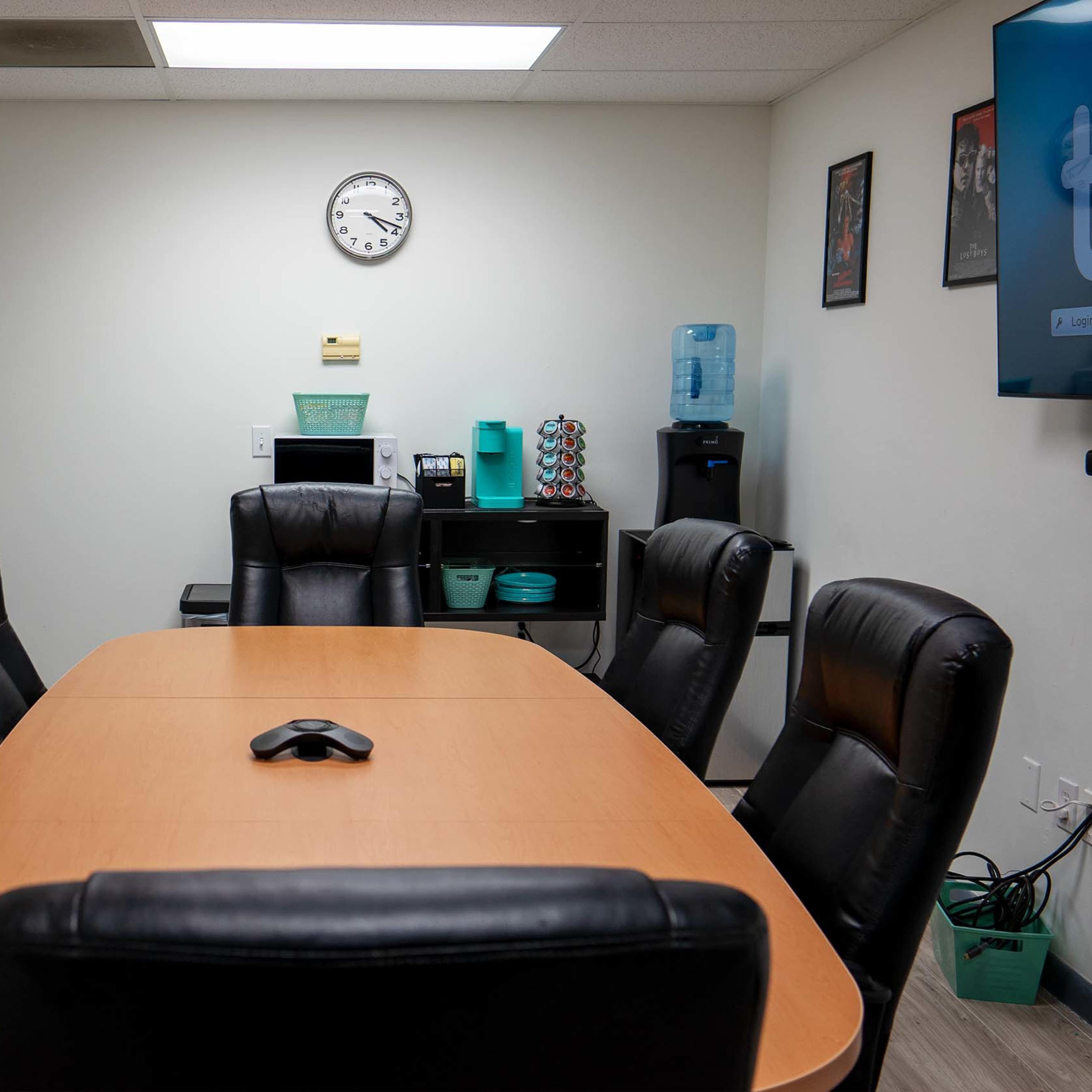A conference room features a large wooden table surrounded by black leather chairs, with a wall clock and a television displaying the word "tivi."