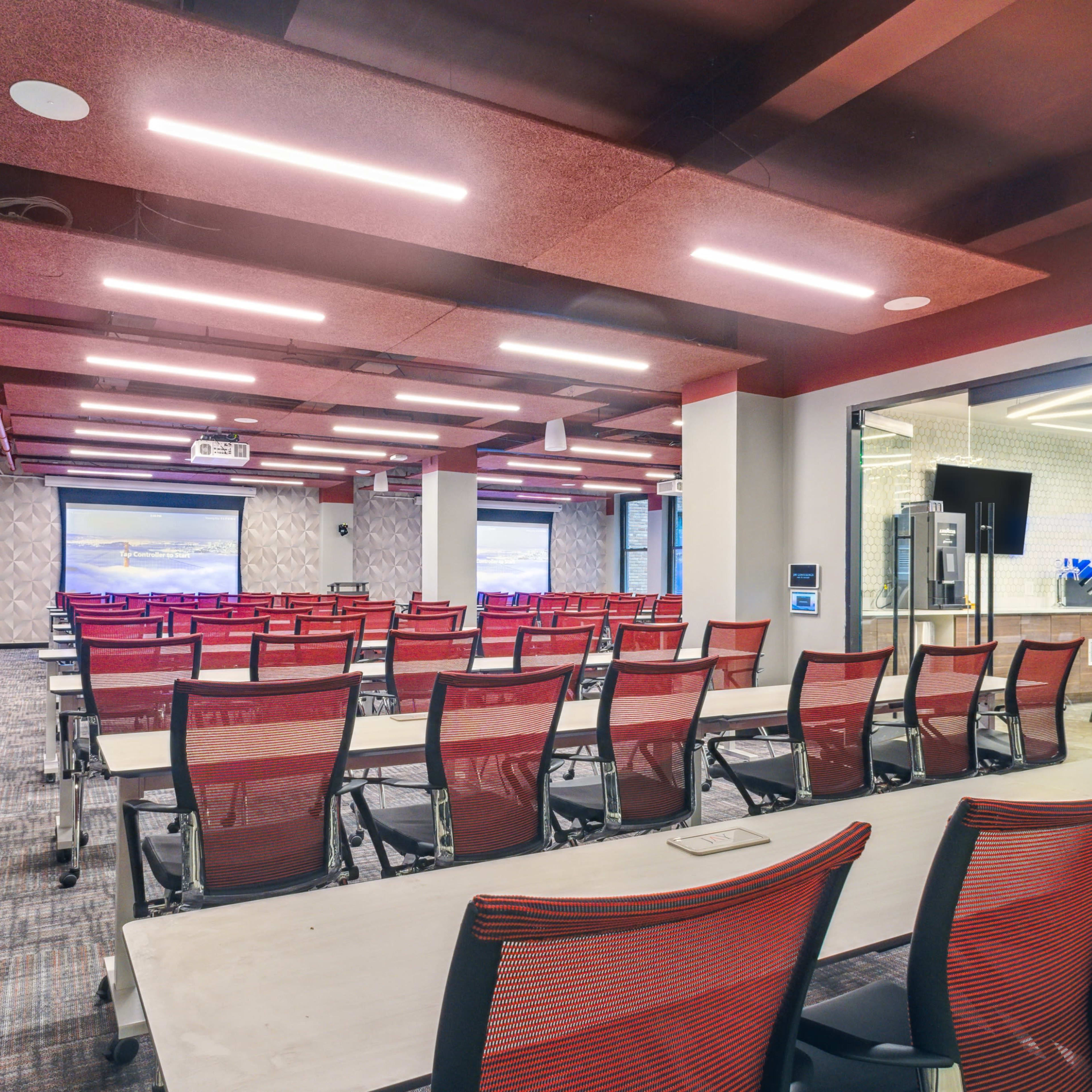 A modern conference room with multiple rows of red and white chairs facing a large screen and two smaller screens on the walls.