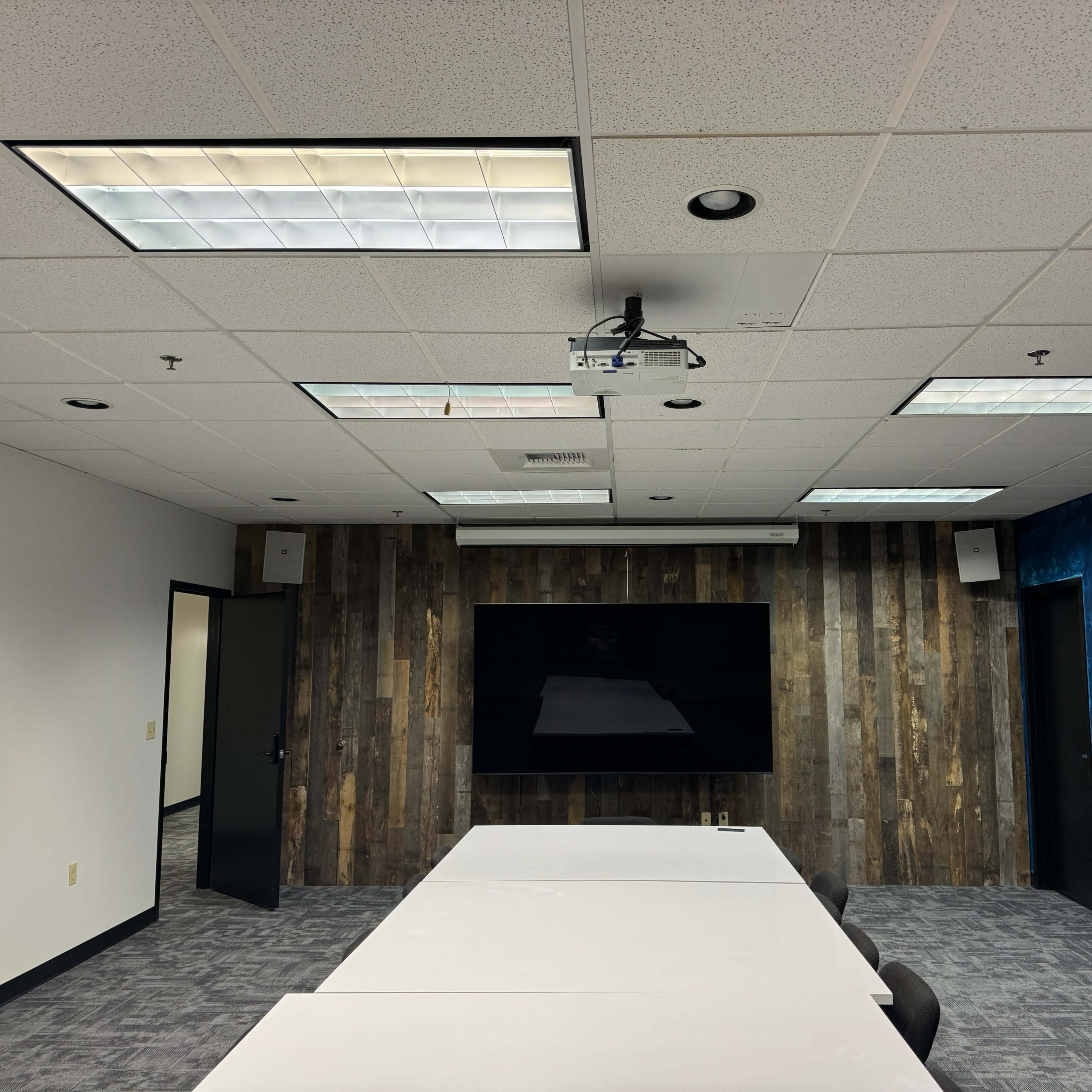 An interior meeting room features a long white table, a large flat-screen TV mounted on a wooden accent wall, and a ceiling-mounted projector.