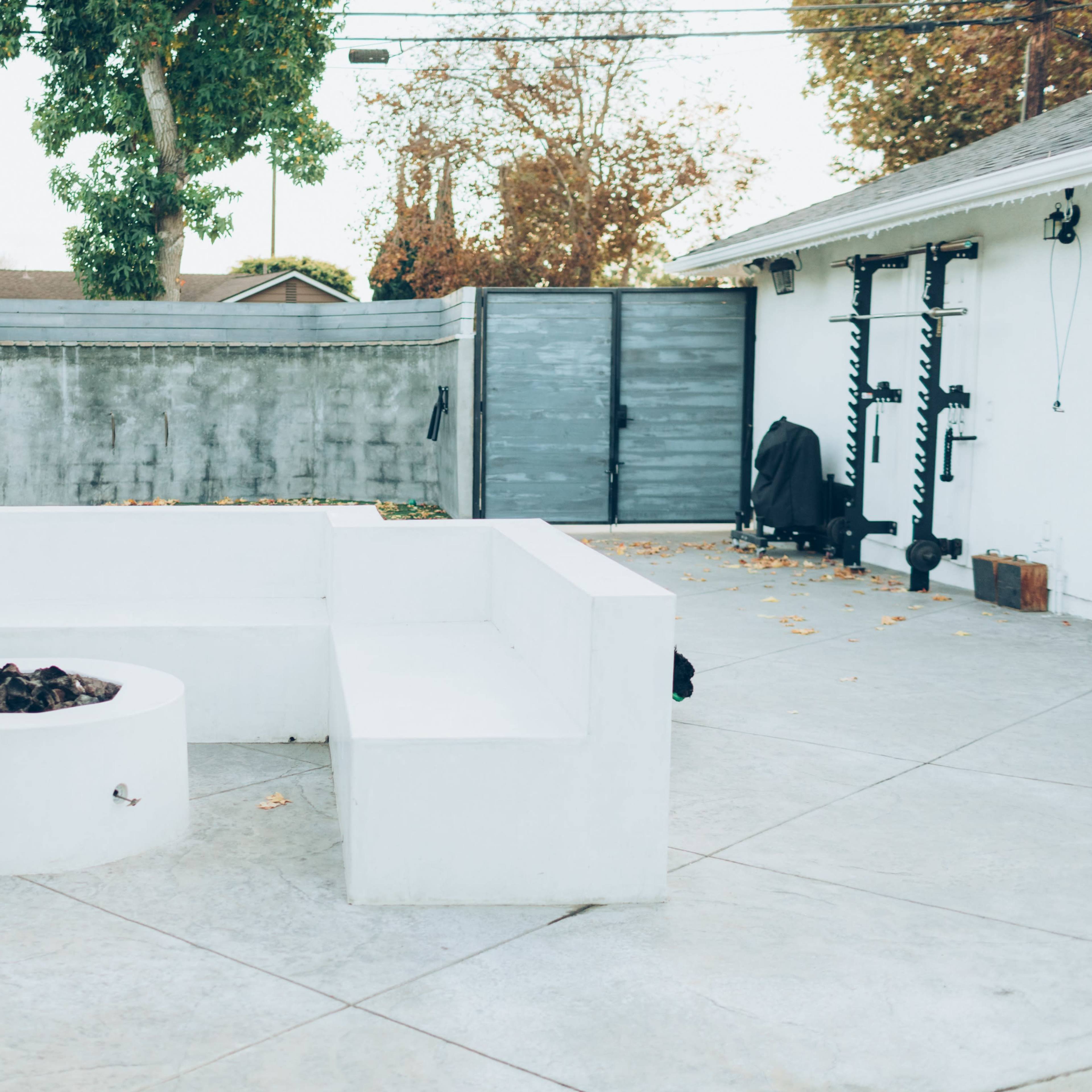 A minimalist outdoor space with a circular fire pit surrounded by a white bench, set against a gray wall and a background of trees.