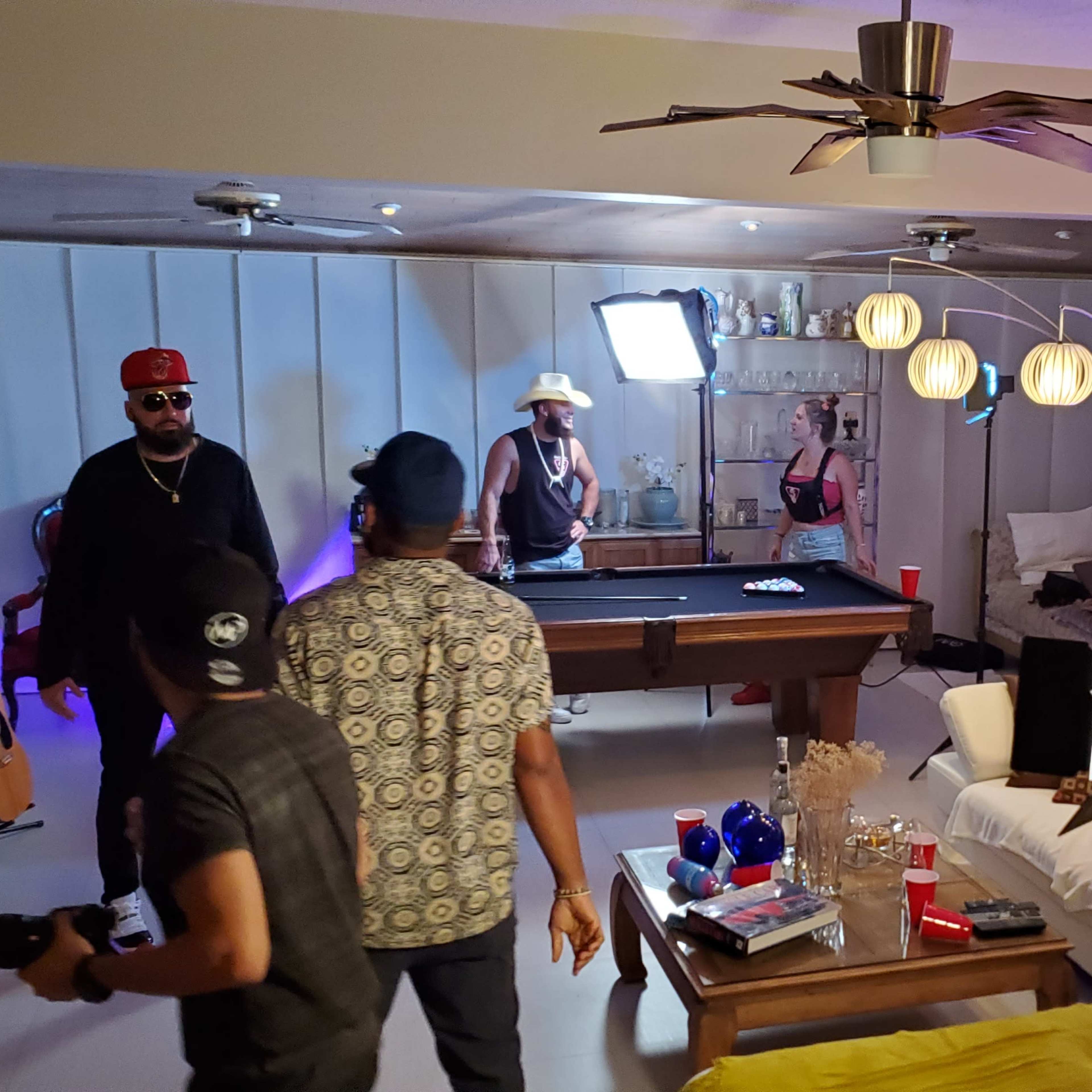 A group of people is gathered around a pool table in a well-lit living room, with cameras and equipment set up for a shoot.
