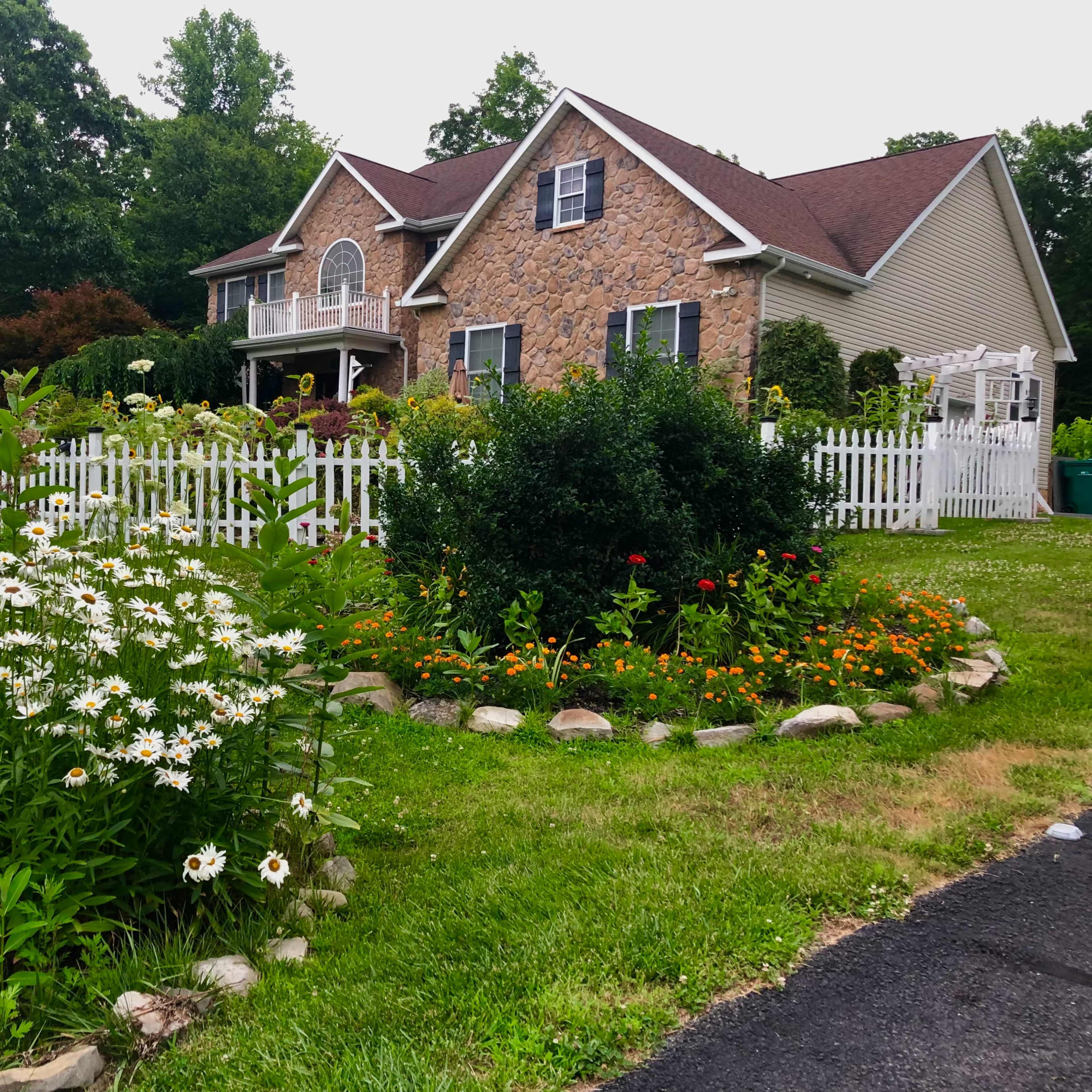 A large two-story house with a stone facade is surrounded by a well-kept garden featuring a circular flower bed and white picket fencing.