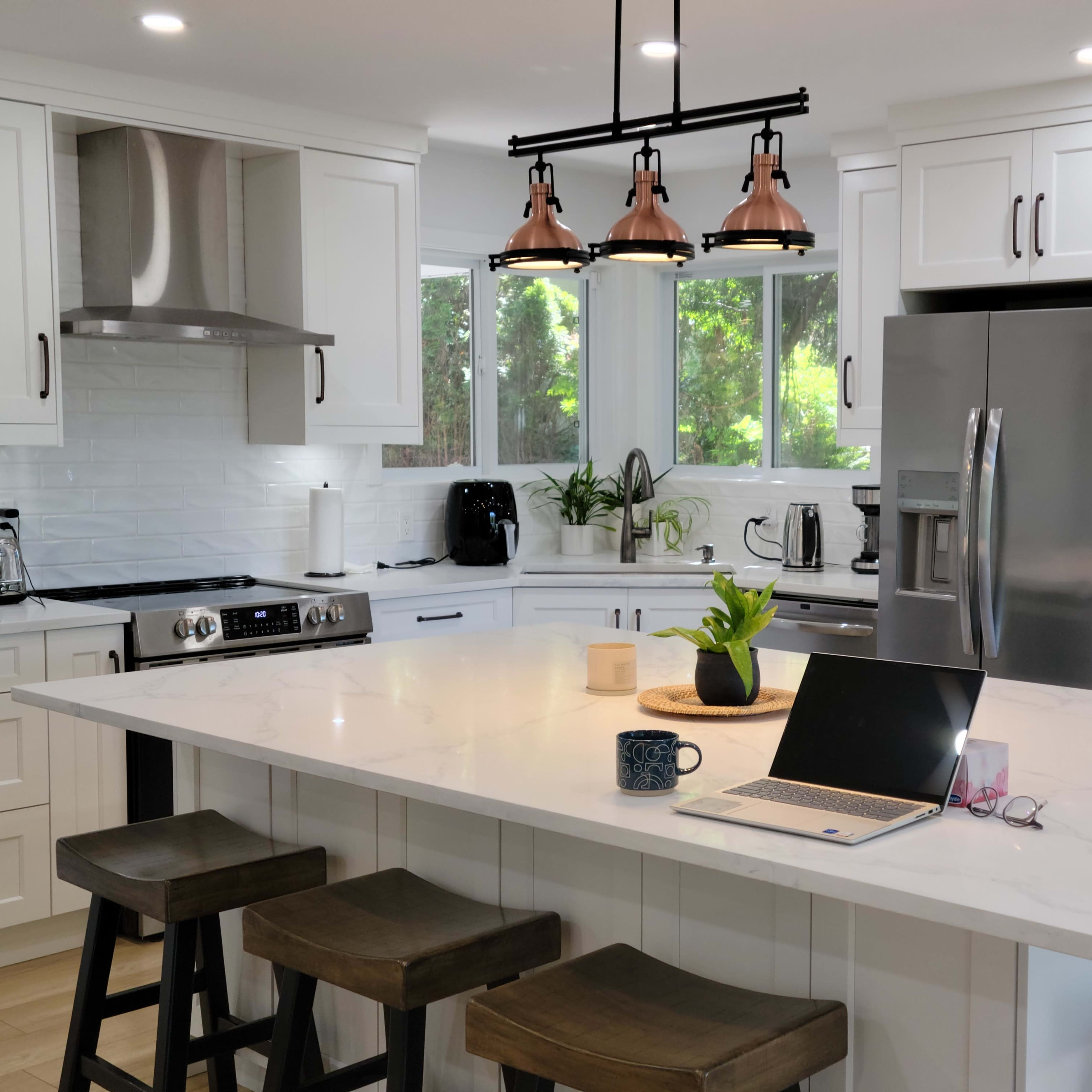 A modern kitchen features white cabinets, a large island with a marble countertop, and a laptop on the surface, surrounded by plants and stainless steel appliances.
