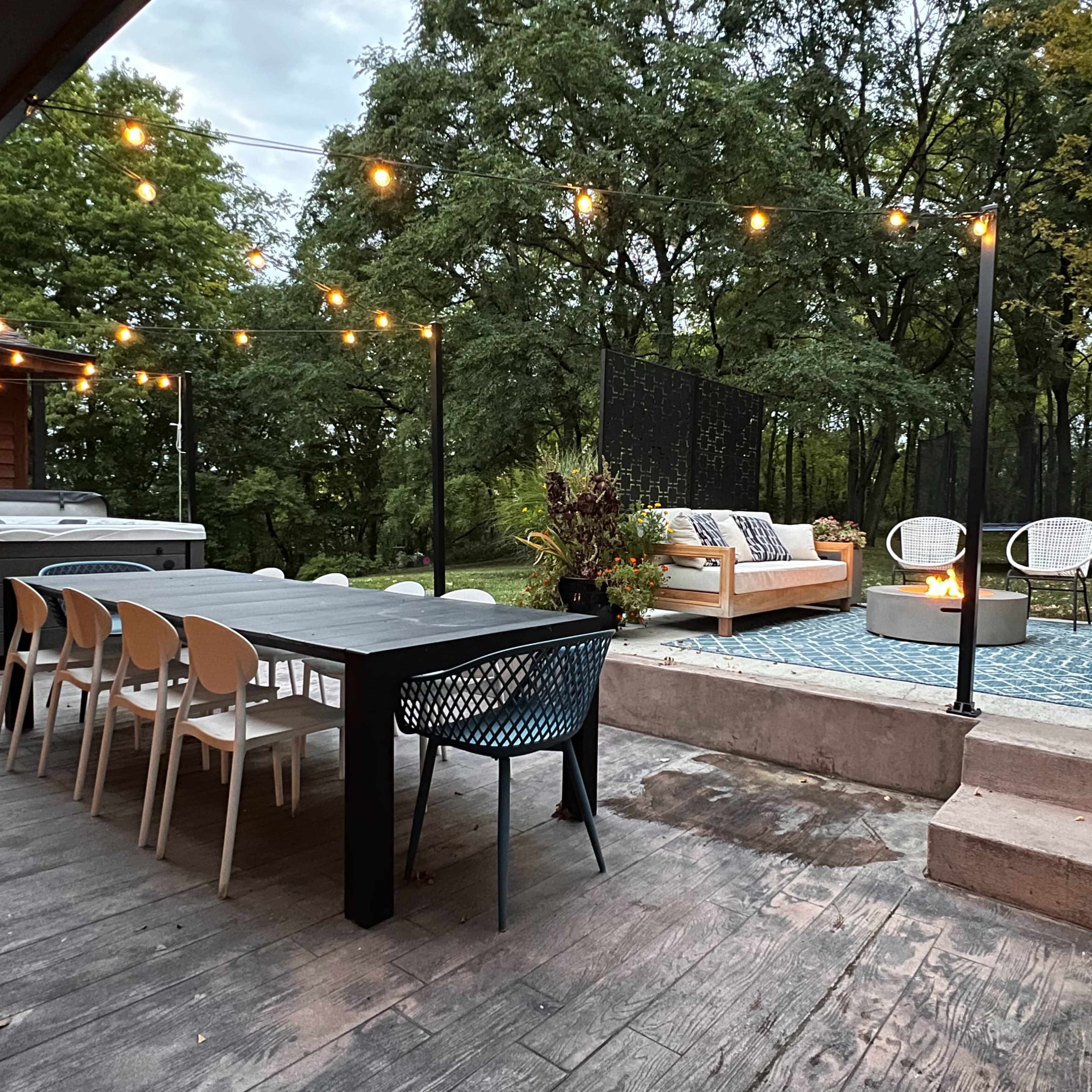 A backyard patio area with a long dining table, outdoor seating, a fire pit, and string lights overhead, surrounded by greenery.