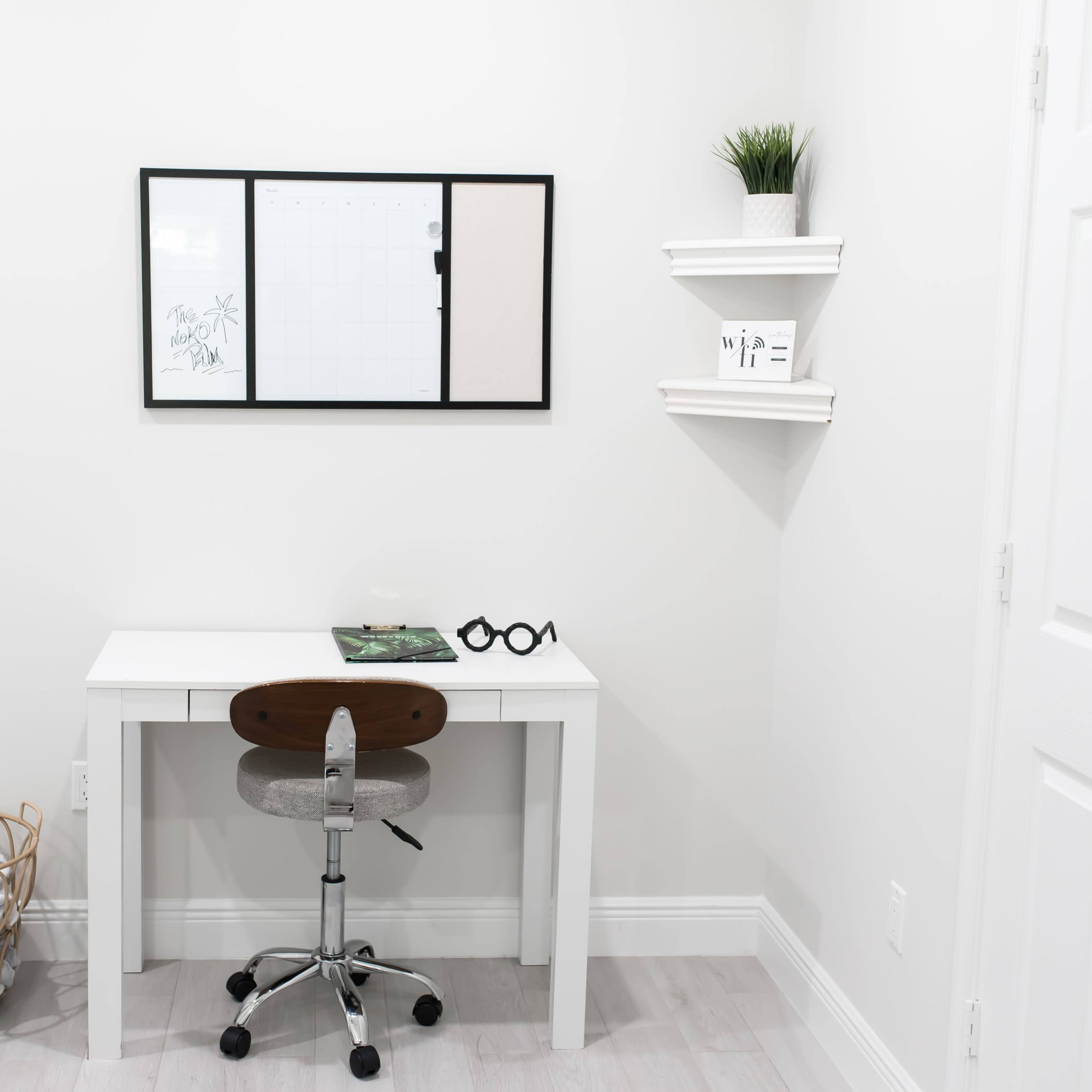A modern, minimalist home office features a white desk with a rolling chair, a wall-mounted grid board above it, and a small shelf holding a potted plant and decorative items.