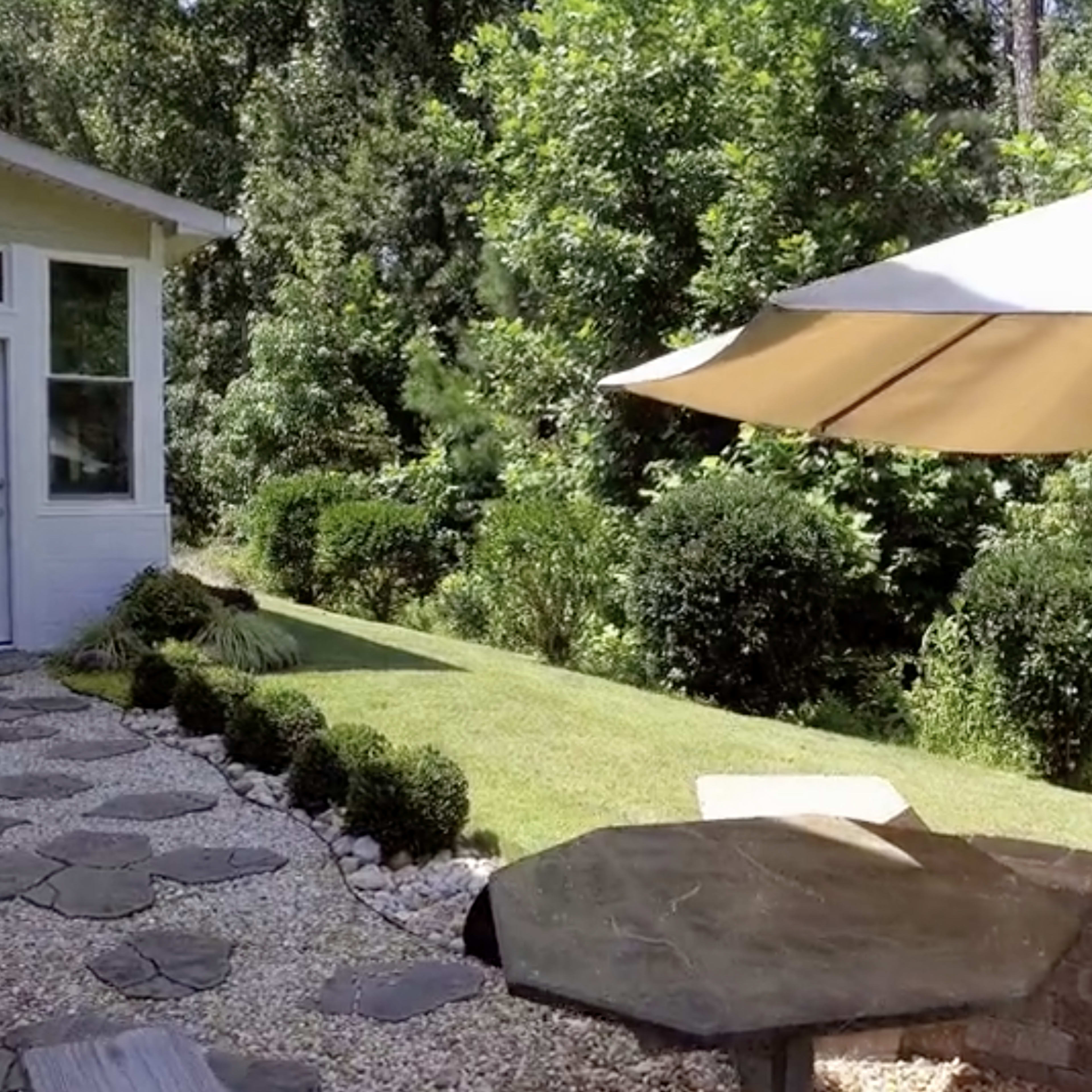A sunlit backyard with a stone pathway leading to a shaded seating area beneath a large umbrella and surrounded by lush greenery.