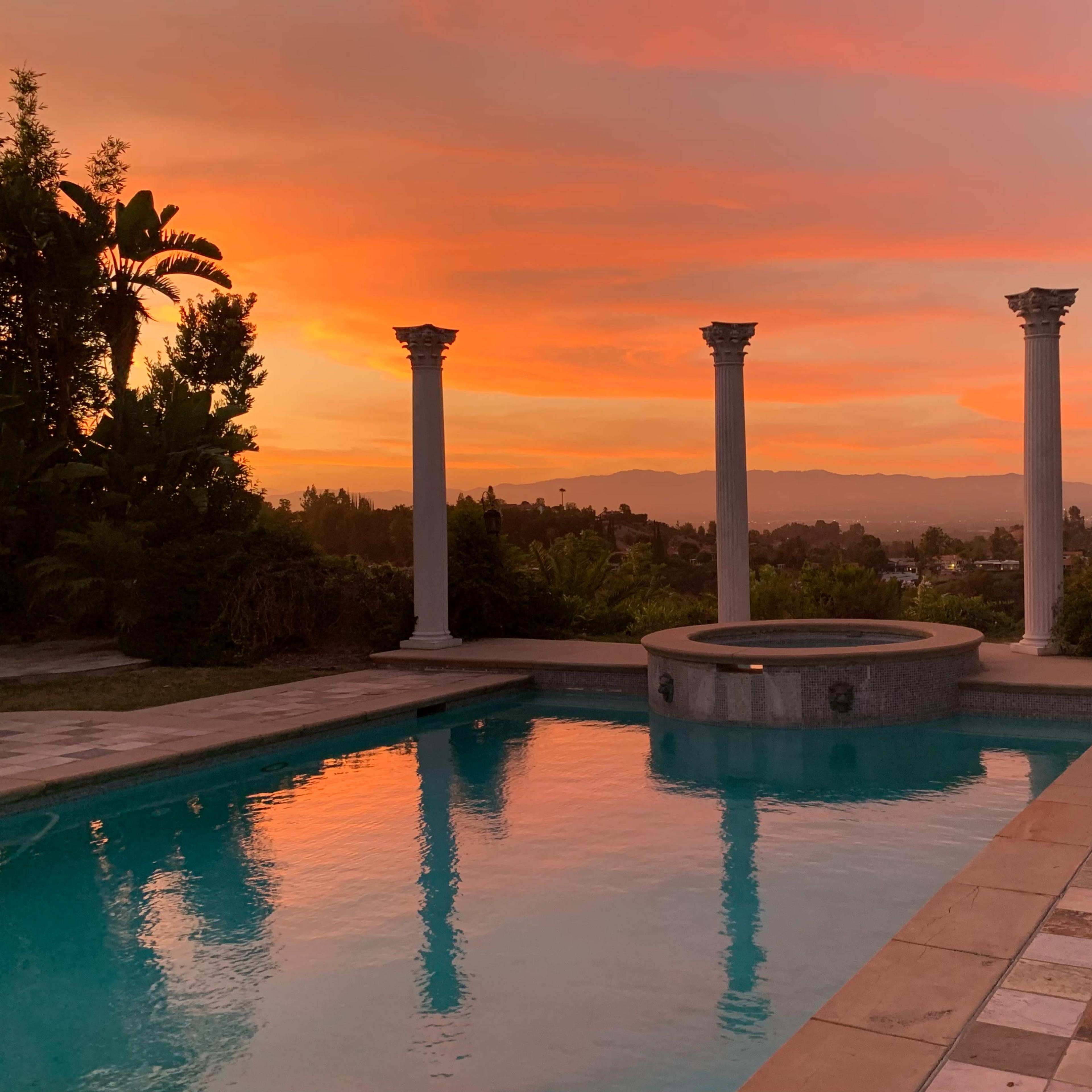 The scene shows a swimming pool with a stone patio, surrounded by tall columns and palm trees, against a sunset sky filled with pink and orange hues.