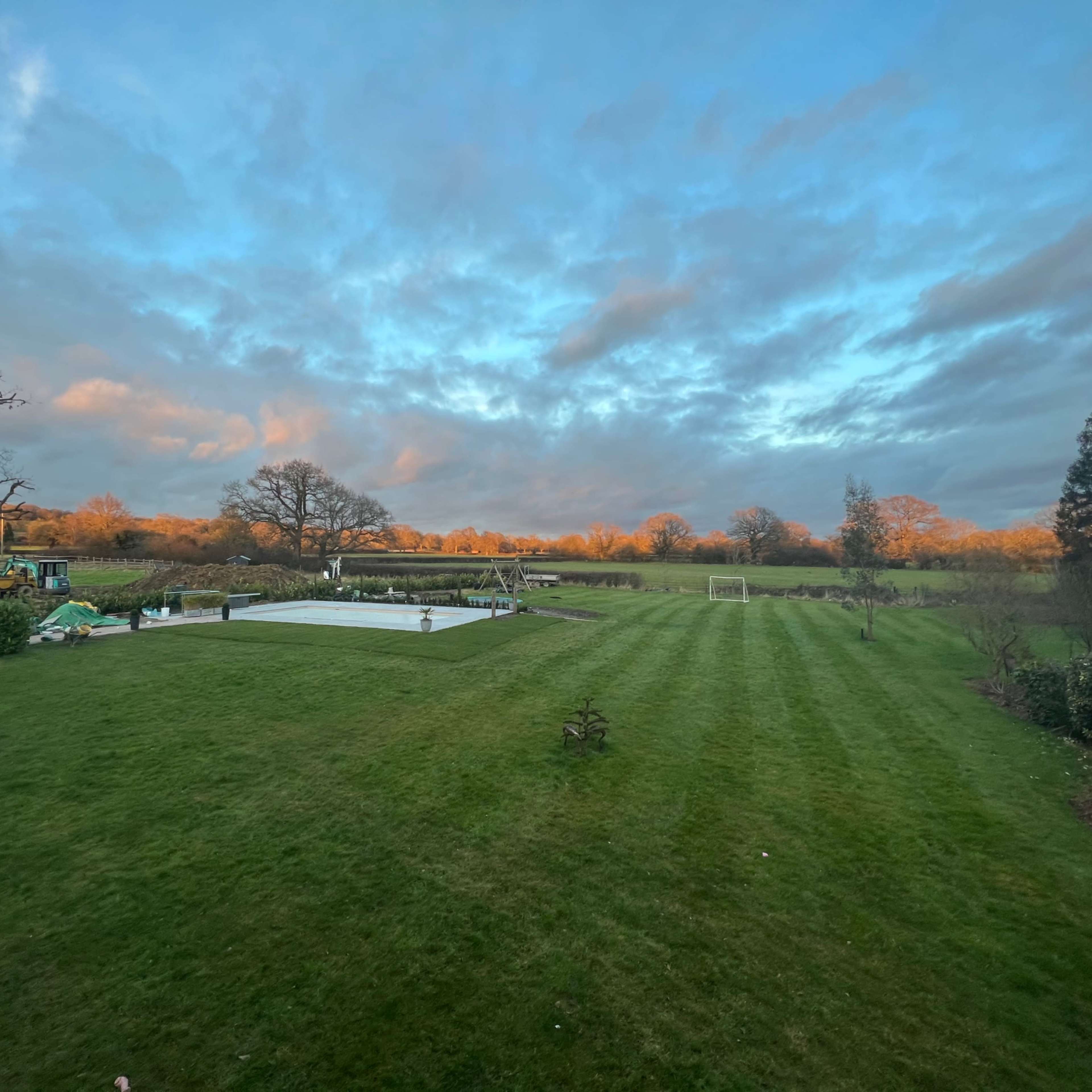 A large green lawn with a covered swimming pool and a soccer goal, set against a backdrop of trees and a cloudy sky.