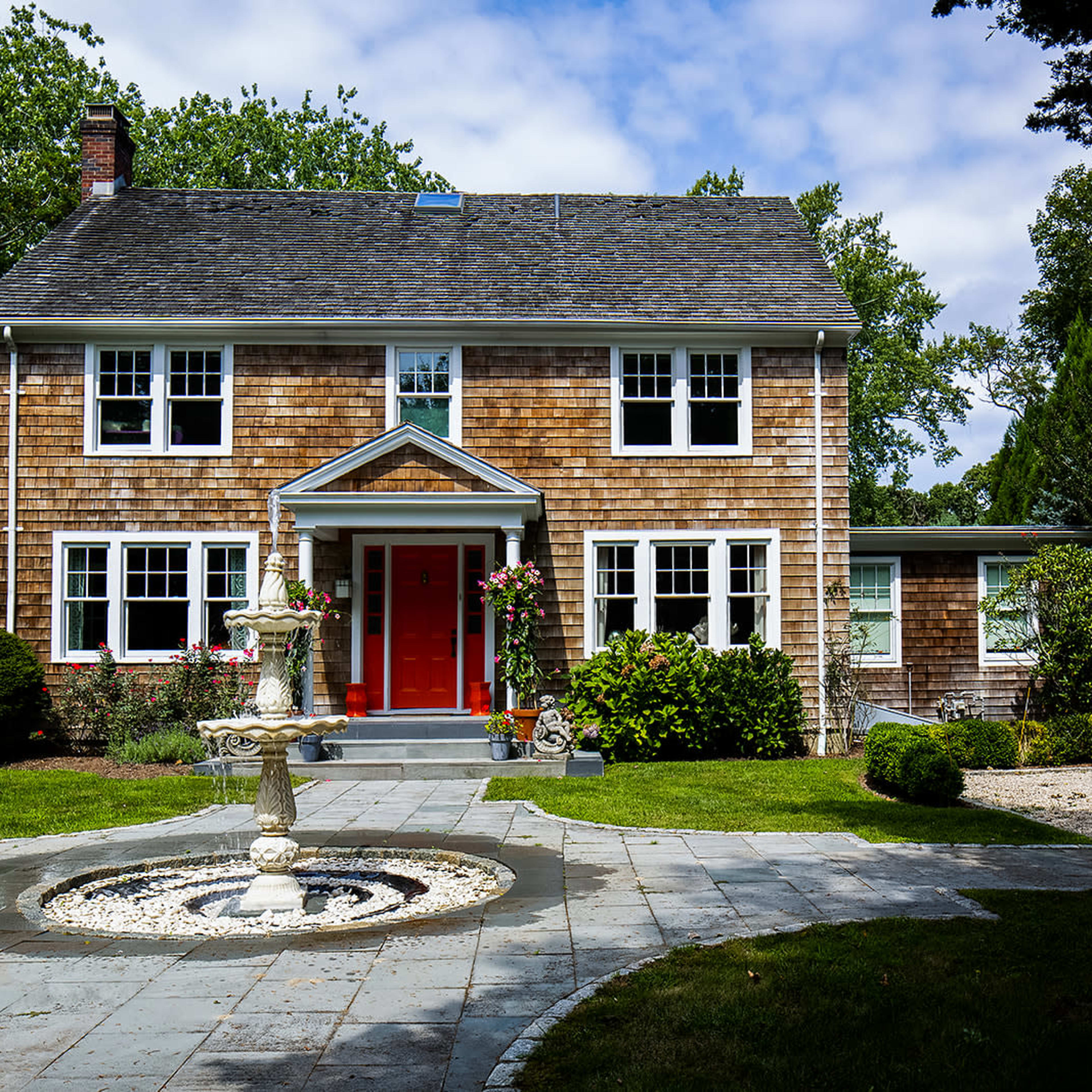 A two-story wooden house with a red front door, surrounded by a landscaped garden and a stone fountain in the foreground.