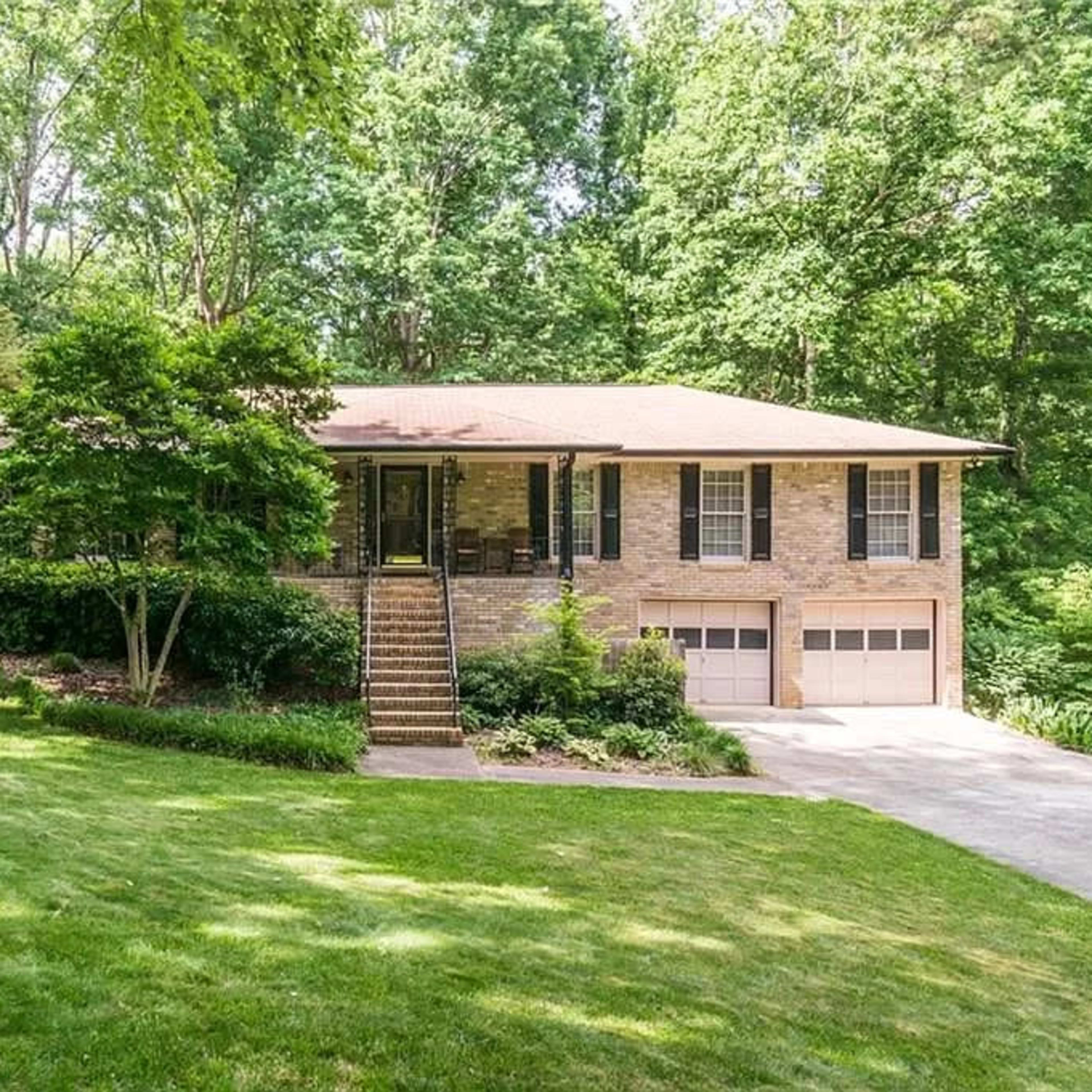 A brick house with a front staircase is set against a backdrop of dense green trees and a well-maintained lawn.