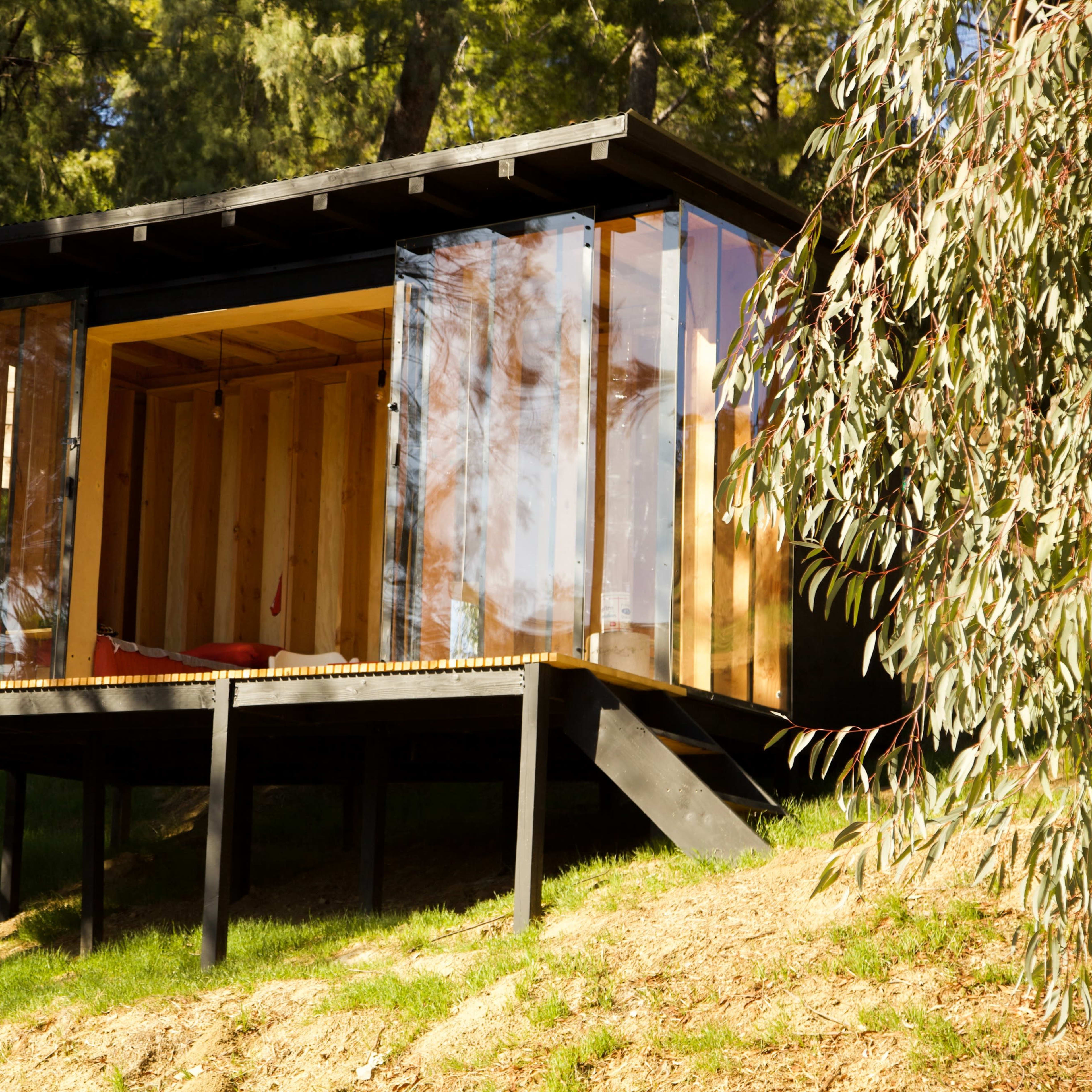A modern glass and wooden cabin sits on a sloped landscape surrounded by trees.
