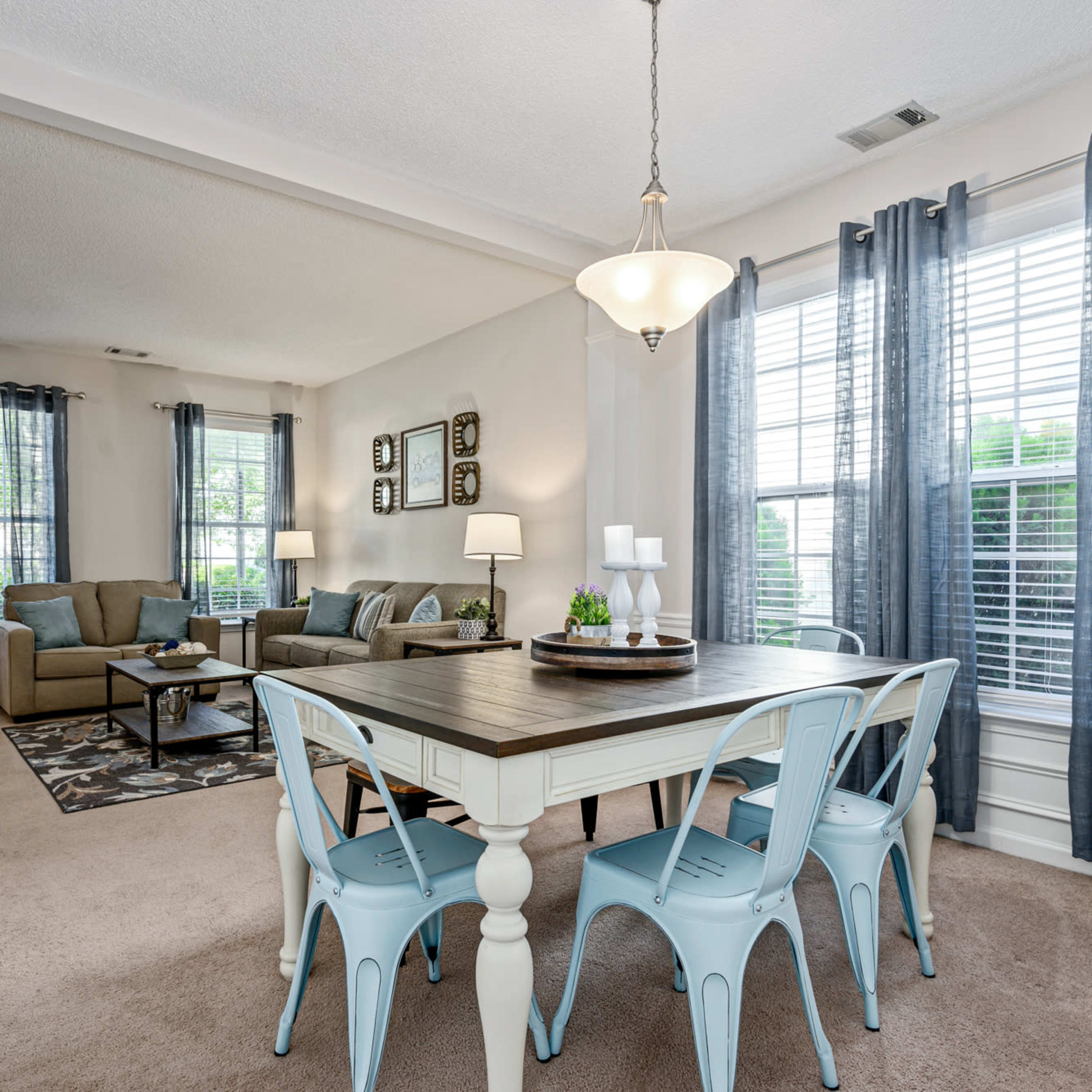 A dining area with a light wooden table and blue metal chairs is situated next to a living space featuring a sofa and coffee table, illuminated by natural light from large windows.