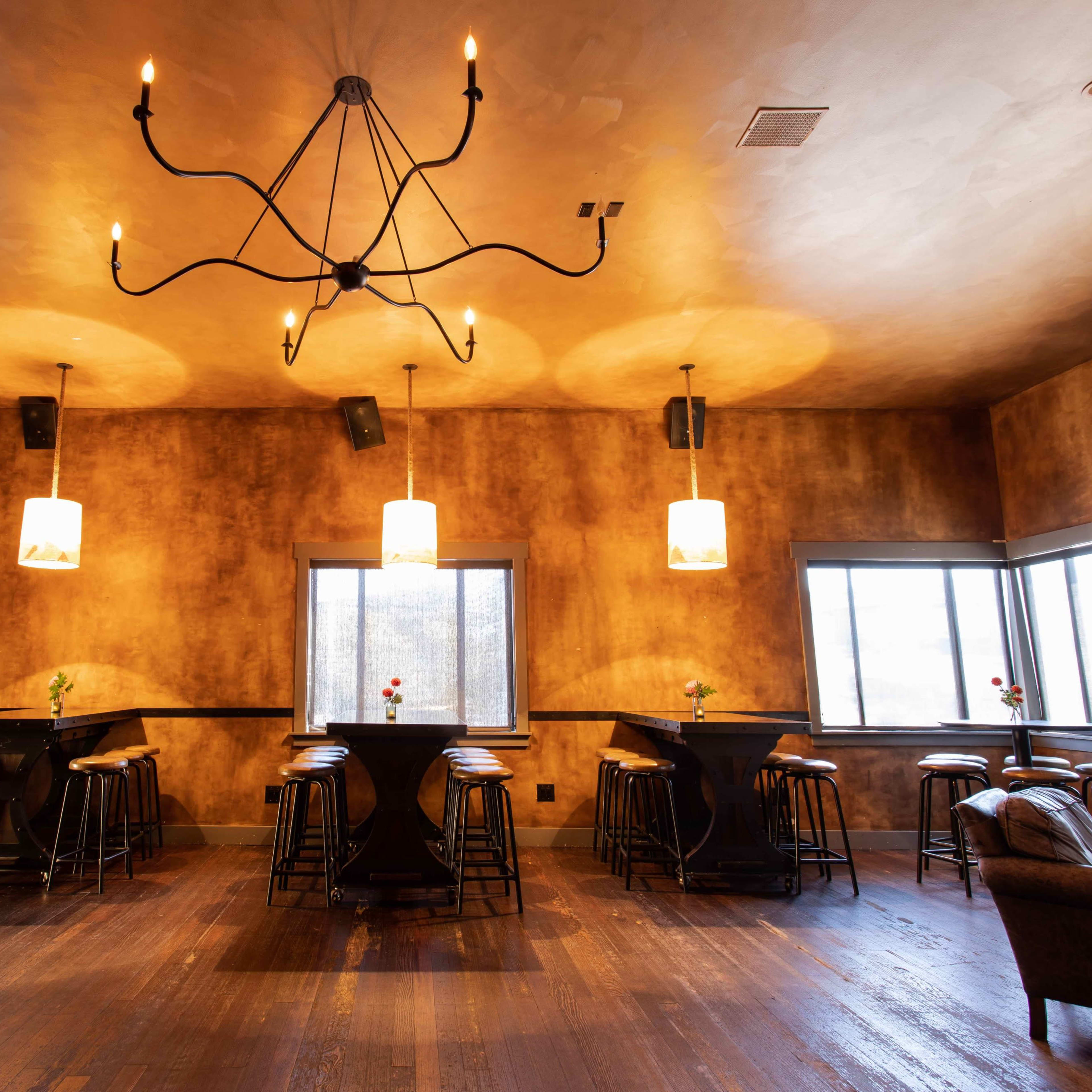 A dimly lit restaurant interior featuring a decorative chandelier, several table setups with stools, and windows allowing for natural light.