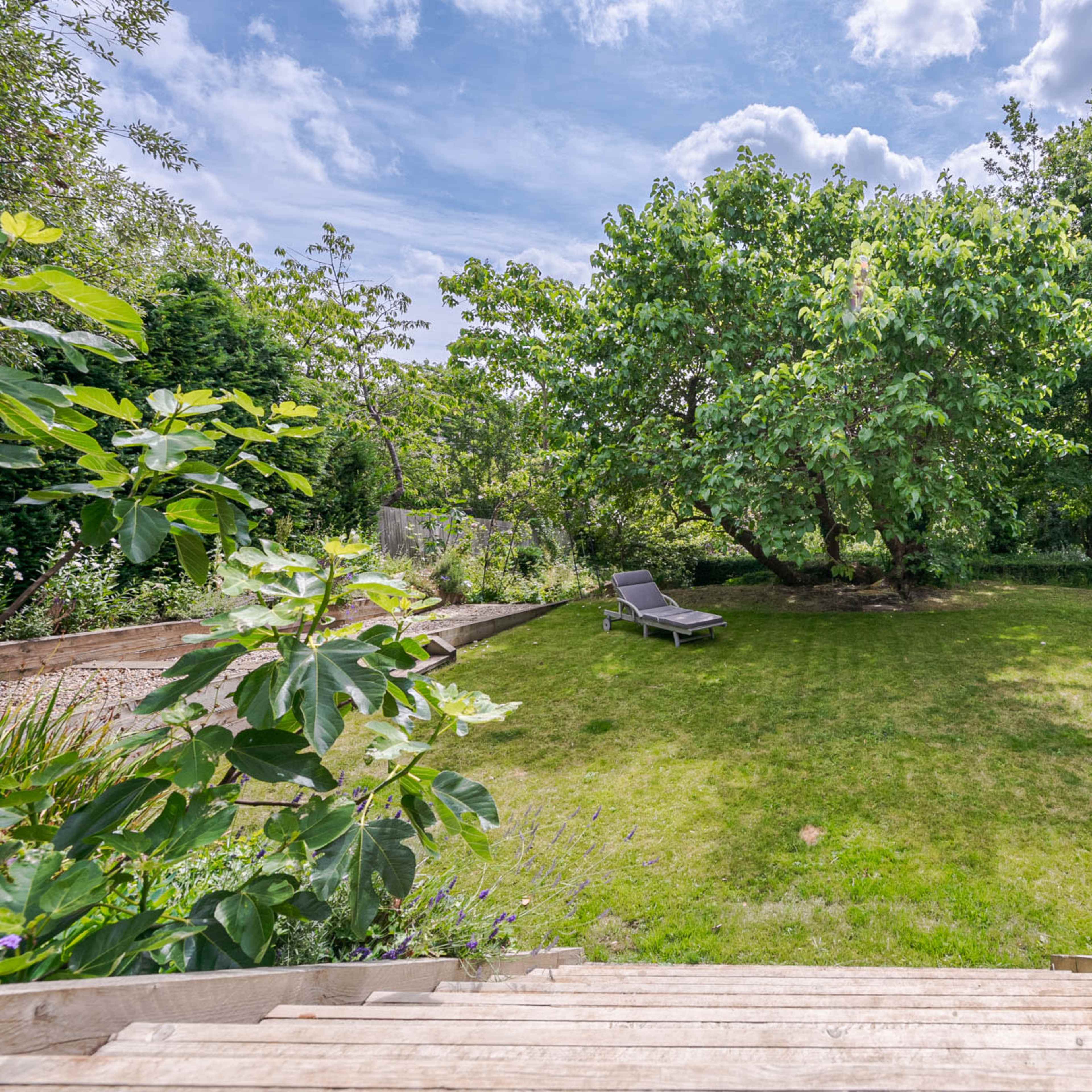 A grassy yard features two large trees and a lounger, surrounded by greenery and flowers.