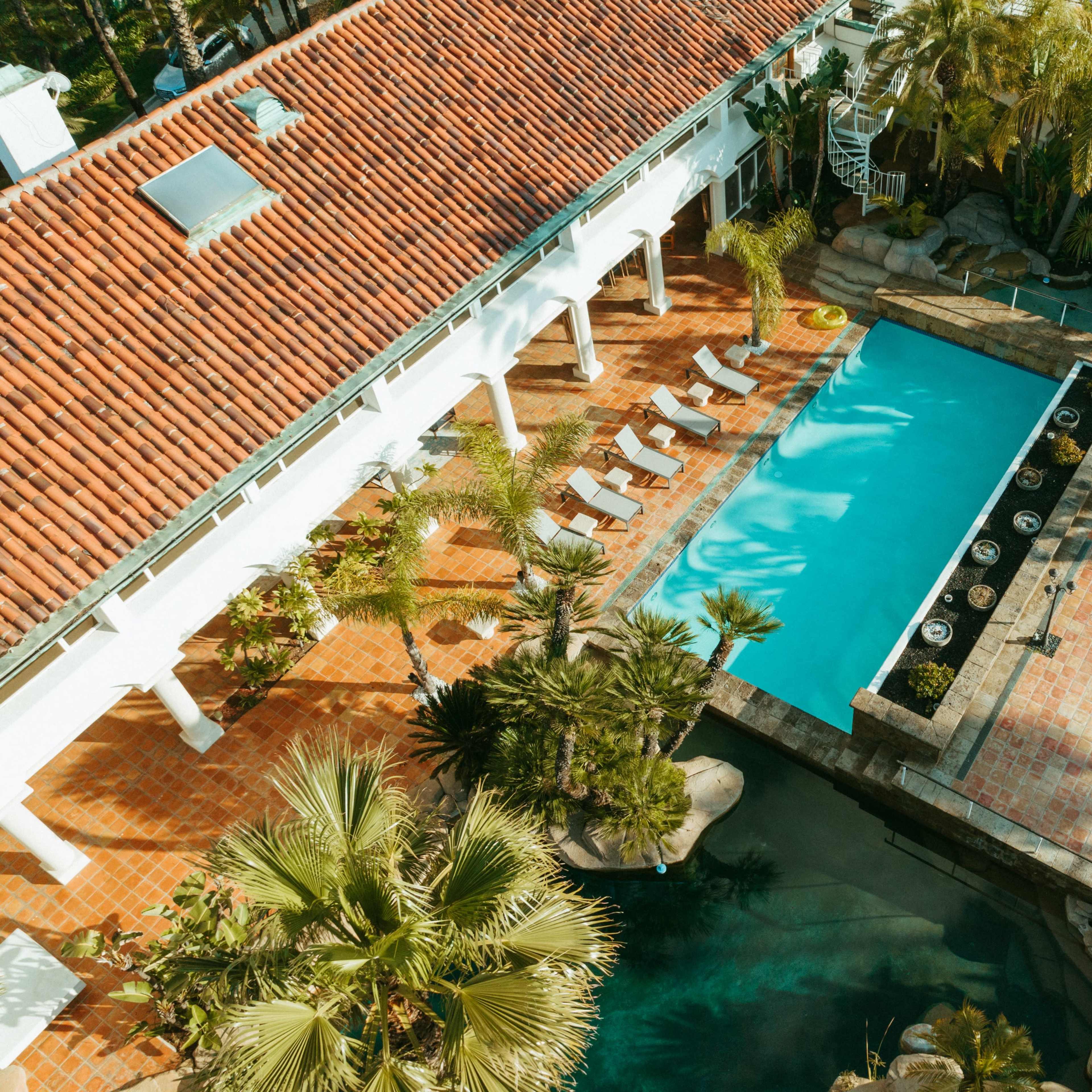 An aerial view of a property with a rectangular swimming pool, surrounded by lounge chairs, tropical plants, and a pathway made of tiled bricks.