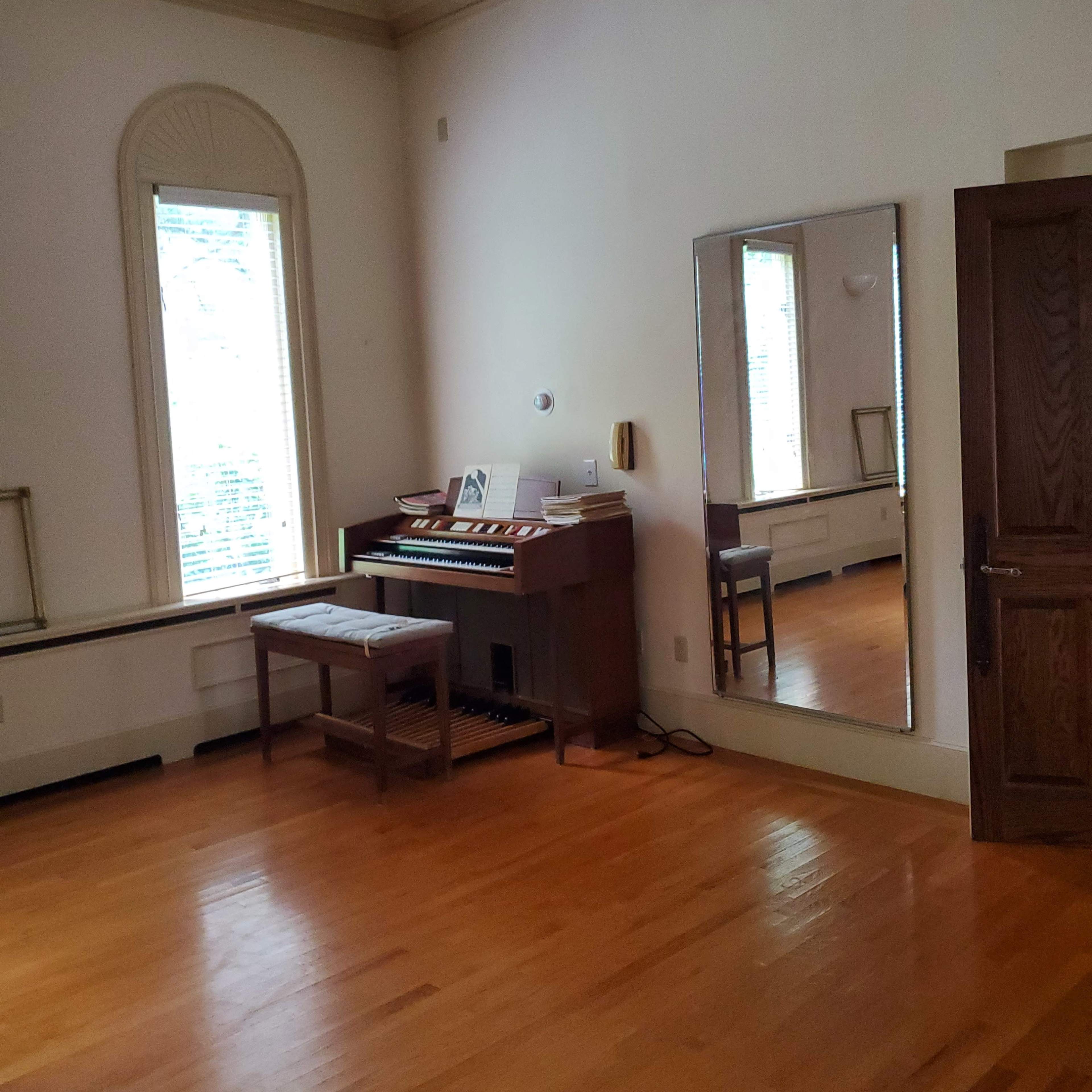 A spacious room with wooden flooring, a piano against the wall, a mirror, and large windows letting in natural light.