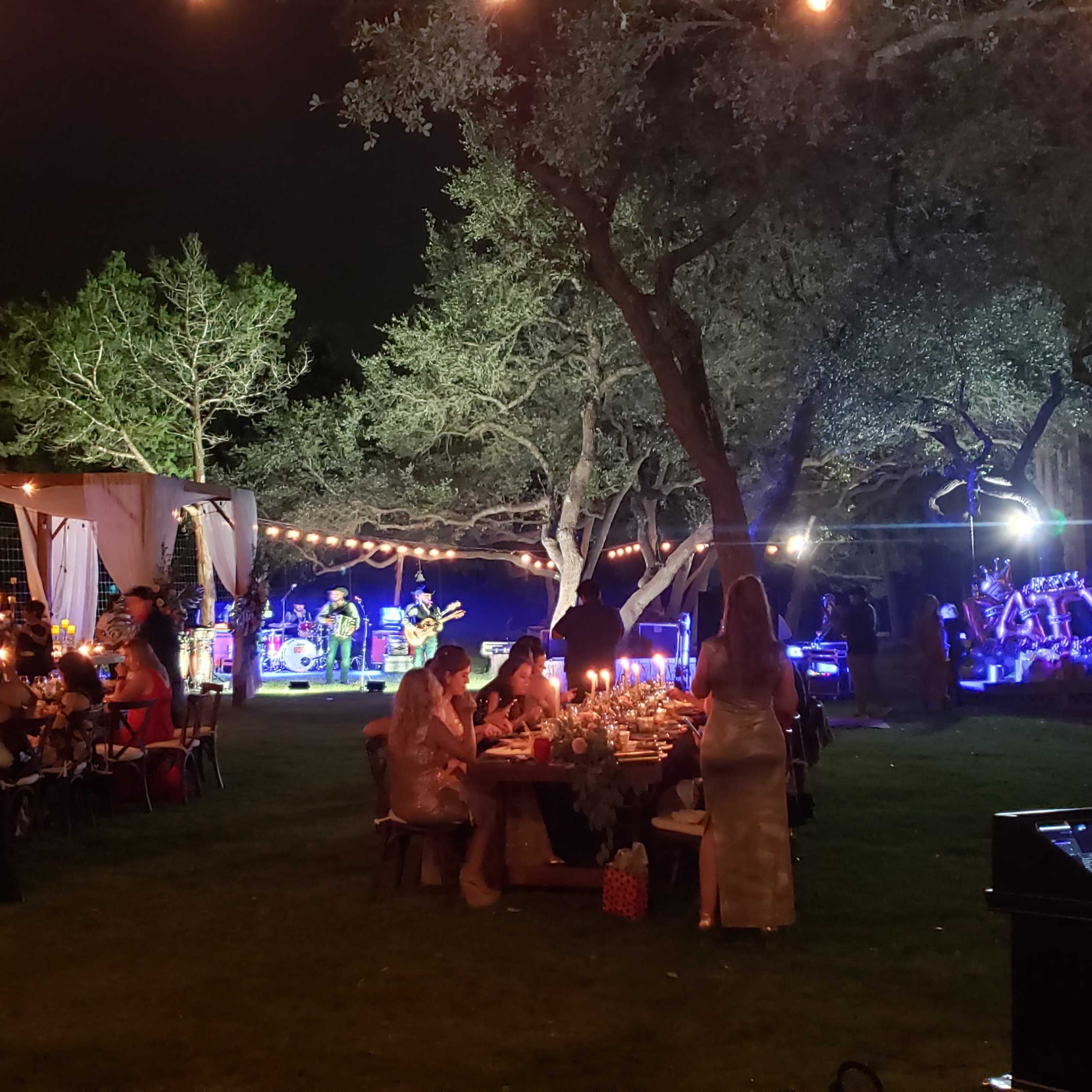 A lively outdoor evening gathering features a band playing music under string lights, surrounded by tables of guests enjoying the festive atmosphere.