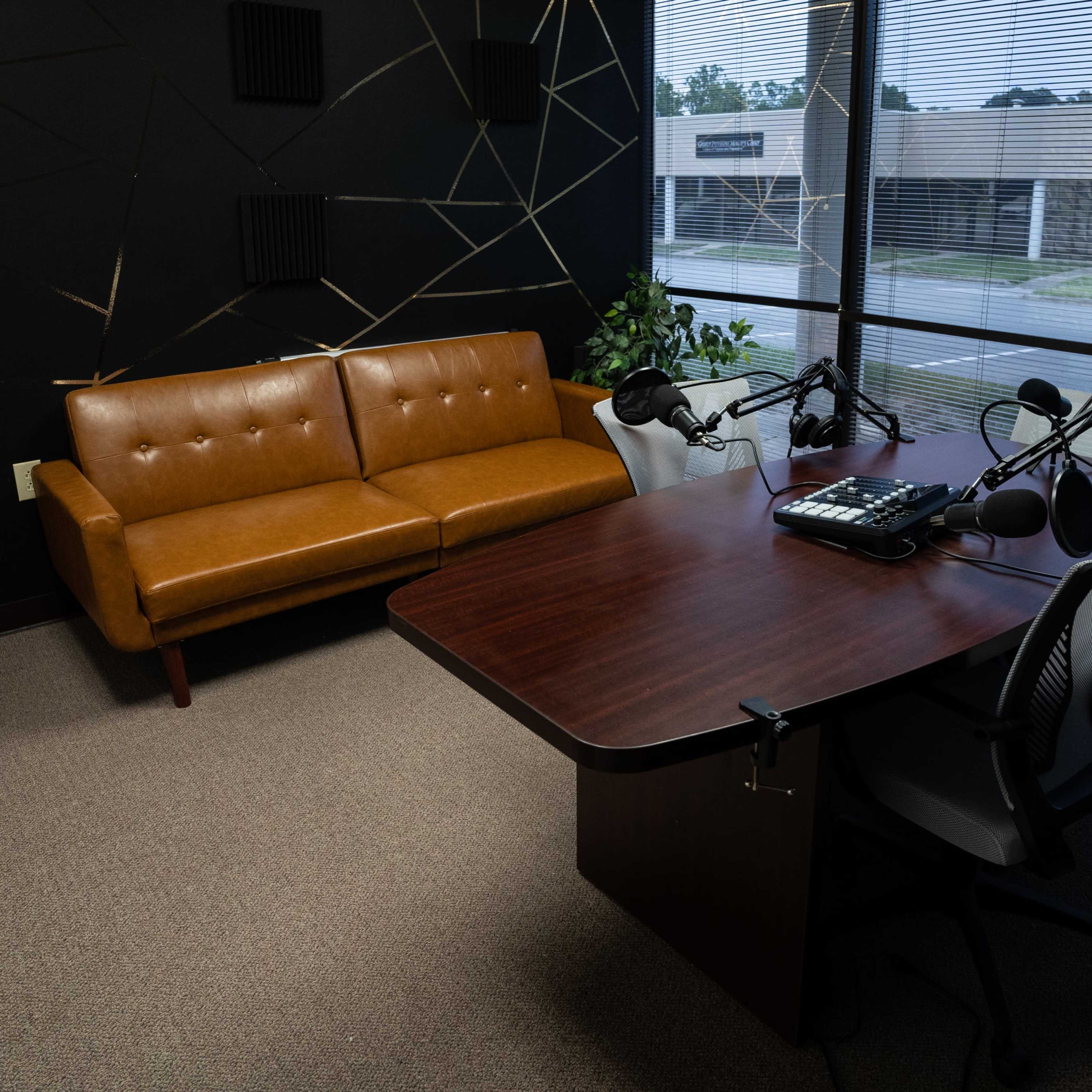 A small recording studio equipped with a brown sofa, a table with microphones, and two office chairs, located near a window with blinds.