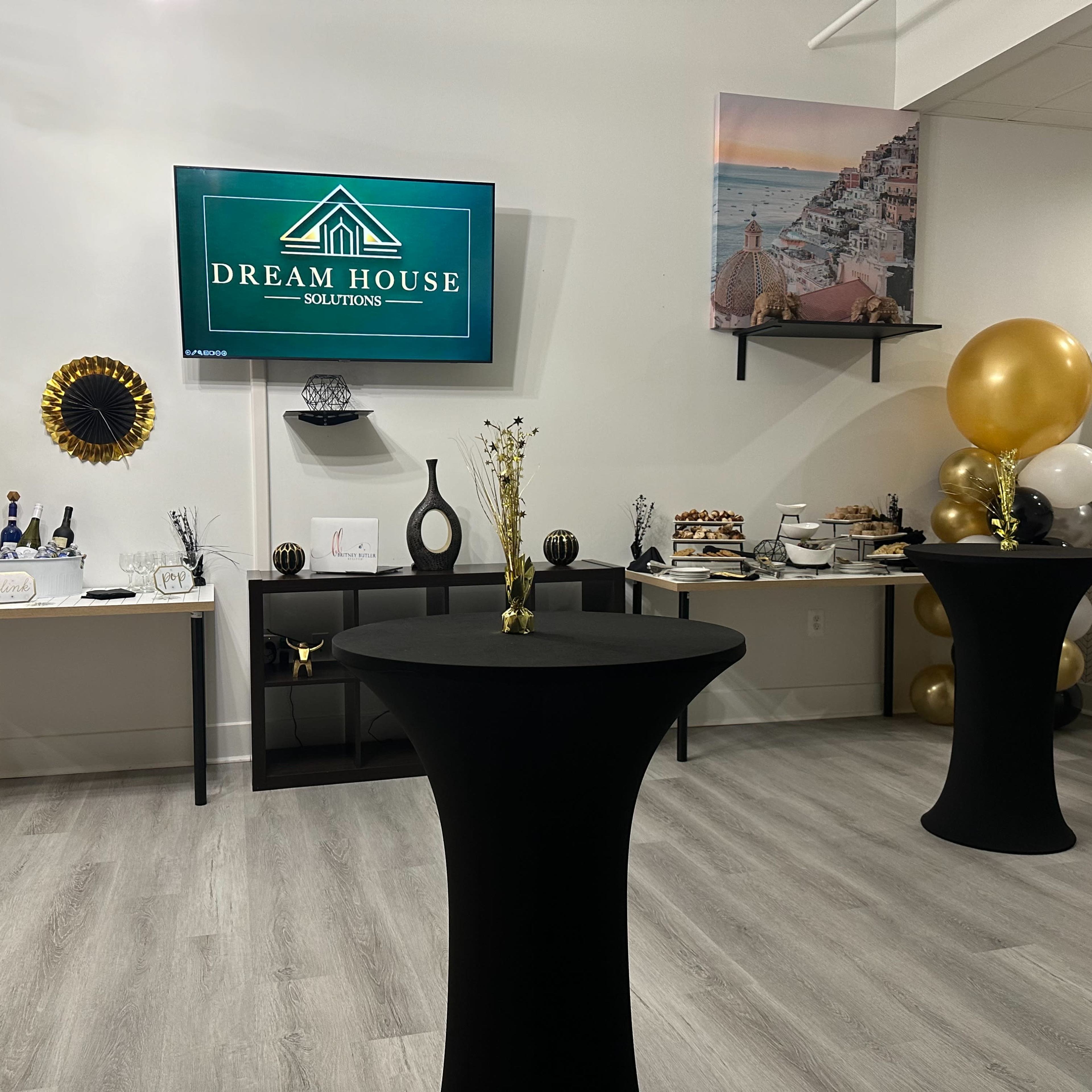 A decorated event space featuring two black tables, a television displaying the "Dream House Solutions" logo, and various food and beverage items on display against a light-colored wall.