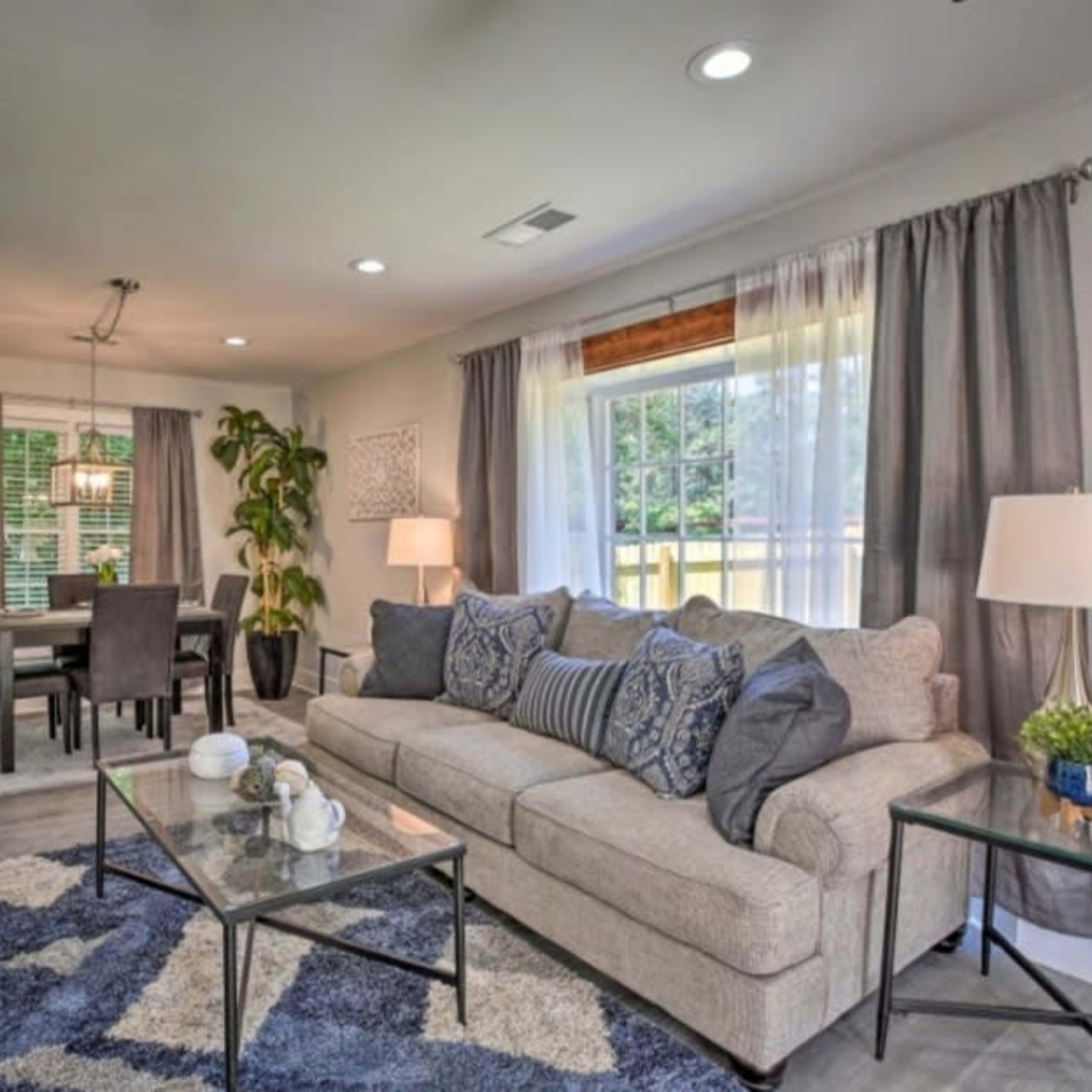 The living room features a light-colored sofa with decorative pillows, a glass coffee table, and a dining area with a chandelier visible in the background.