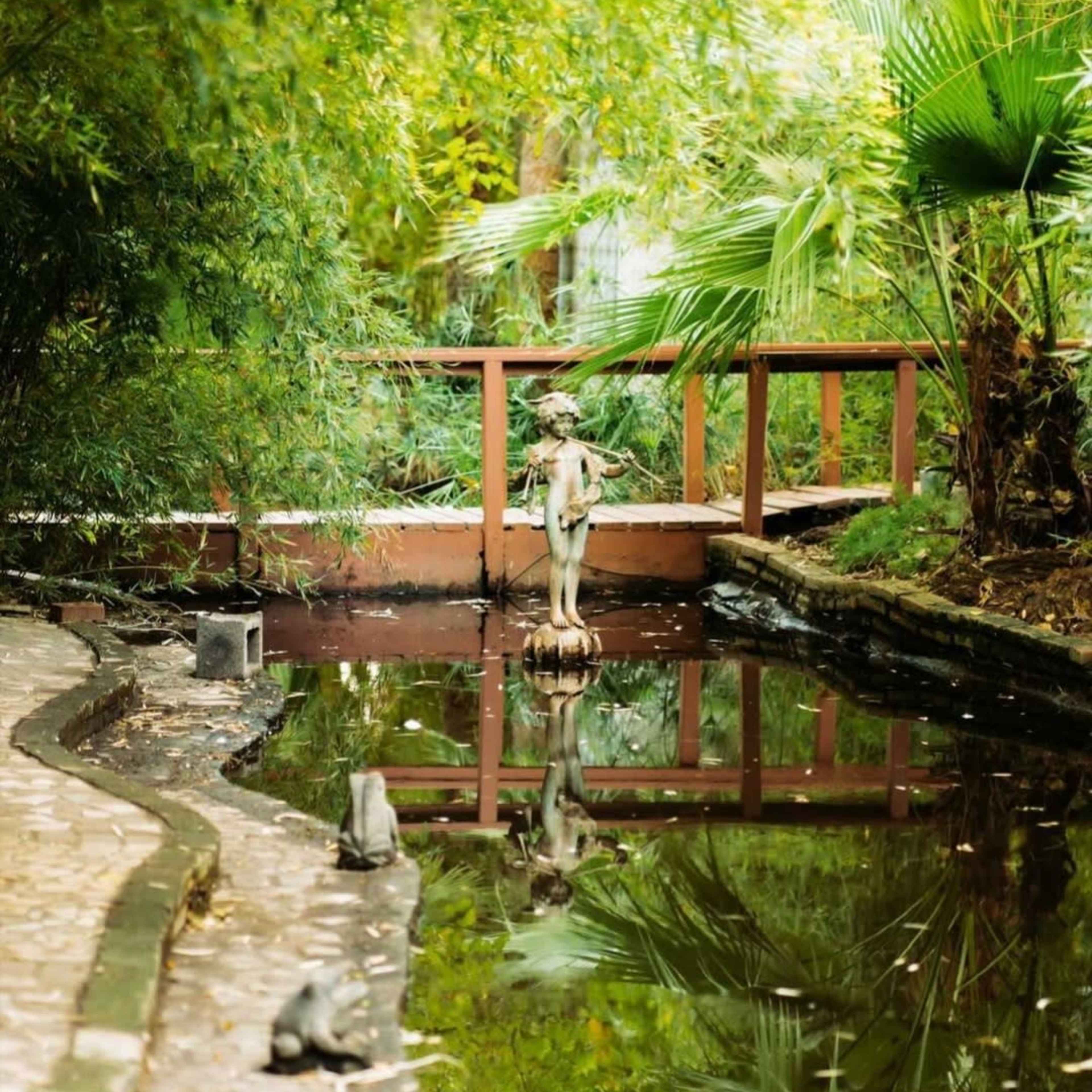 A serene garden path curving alongside a reflective pond, featuring a small wooden bridge and surrounded by lush greenery.