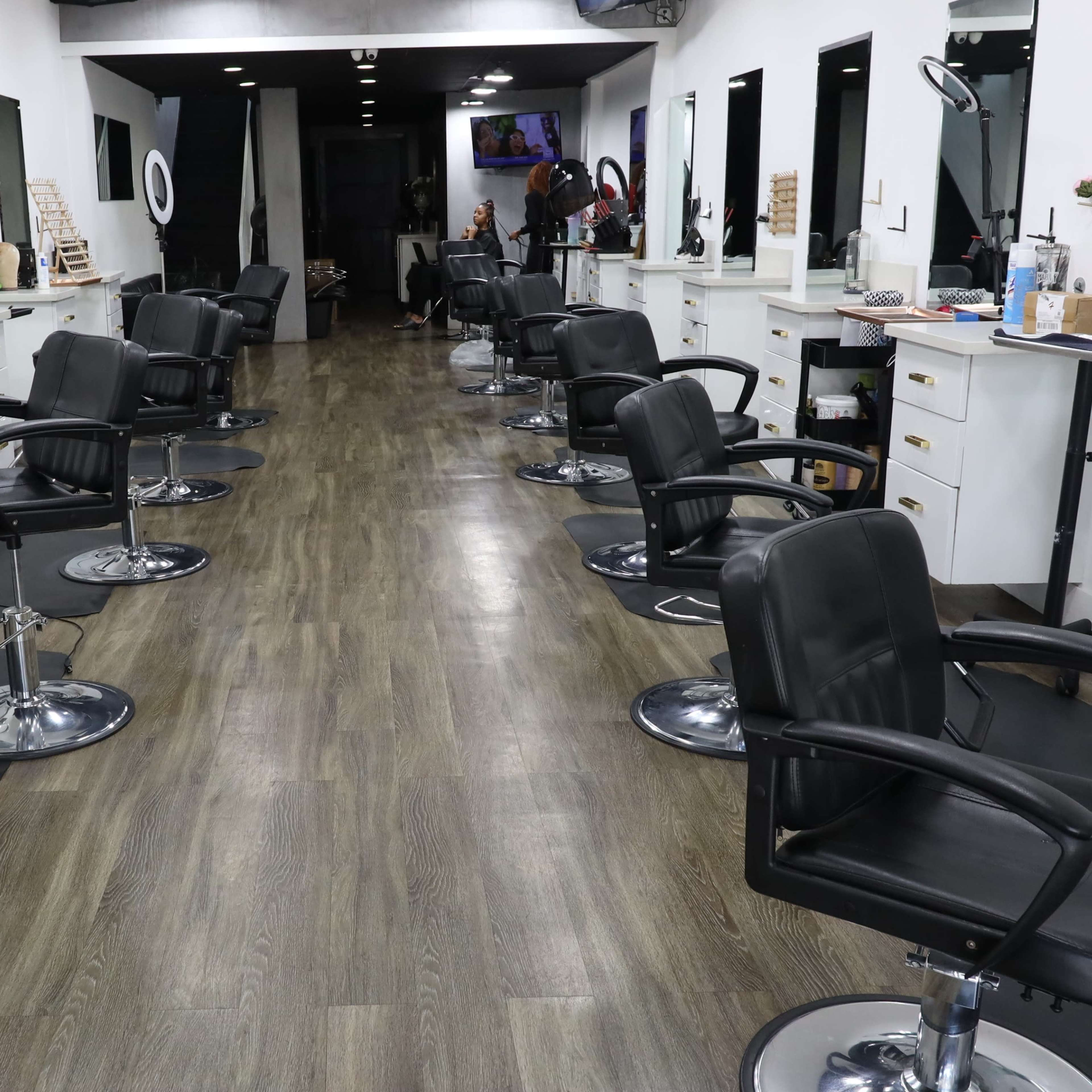A spacious hair salon with several styling stations, each equipped with a black chair and large mirrors, located on either side of a wooden floor aisle.