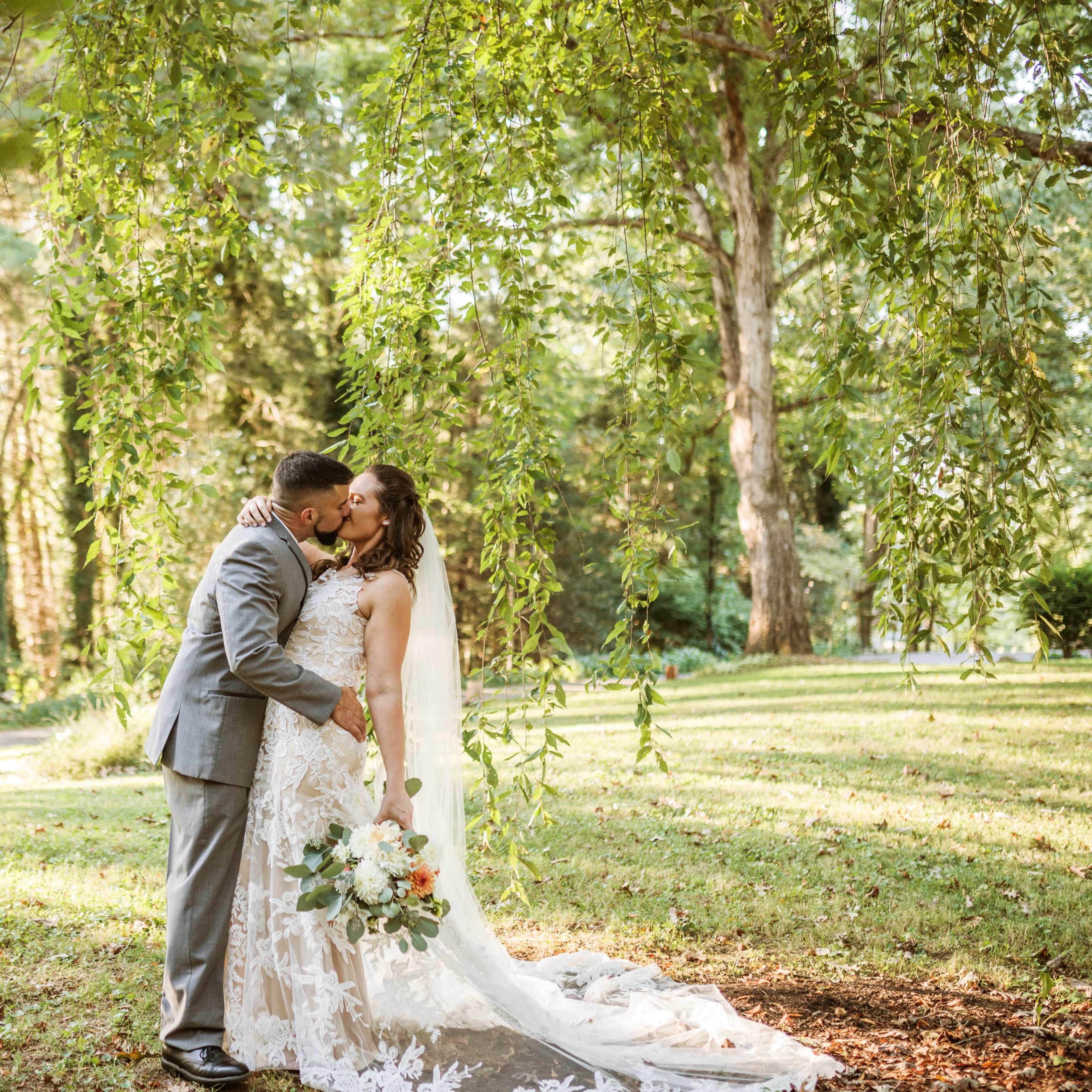 A couple exchanges a kiss under leafy trees in a garden setting during their wedding.