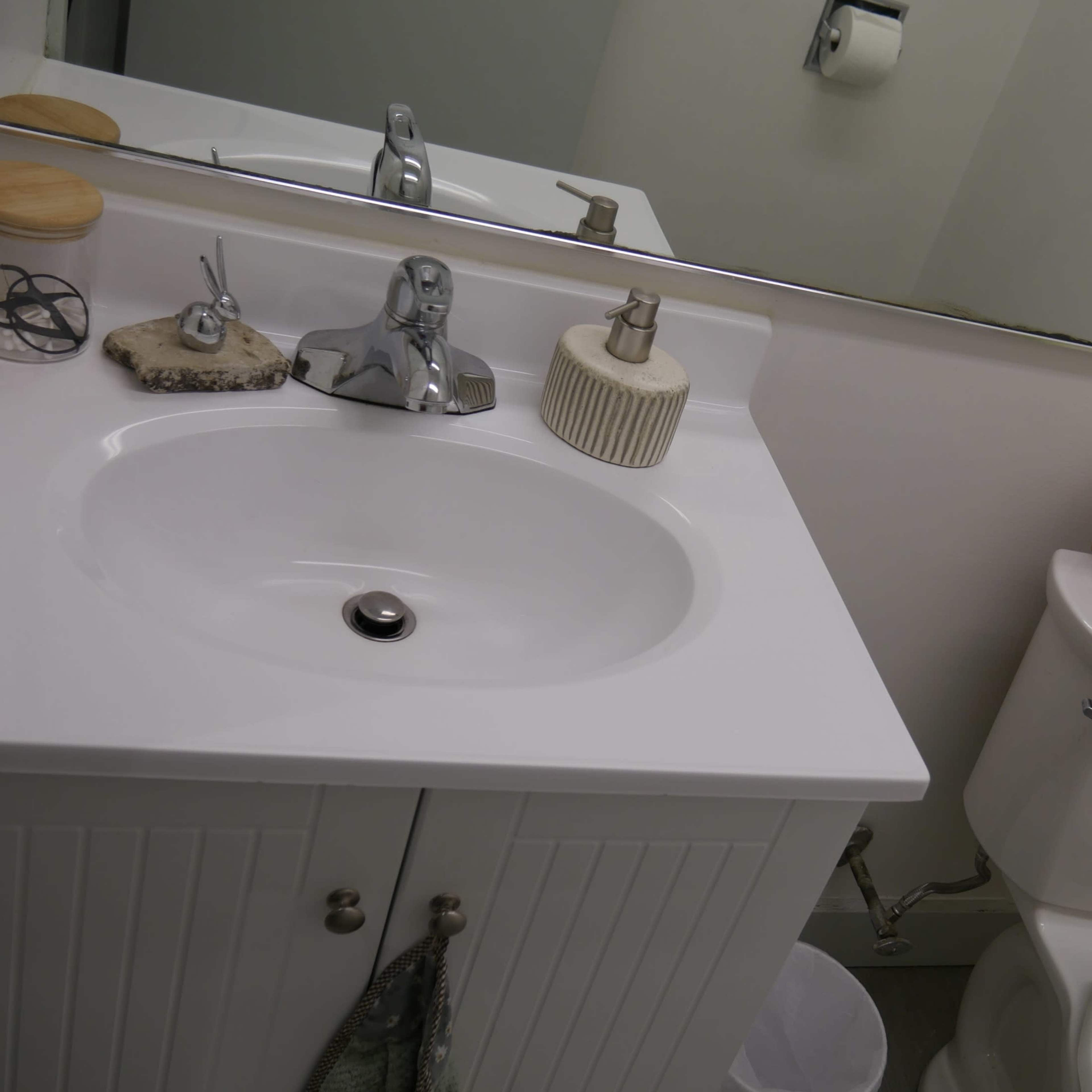 A bathroom sink area with a mirror above it, a faucet, a soap dispenser, and a toilet beside a wicker basket holding toilet paper and other supplies.