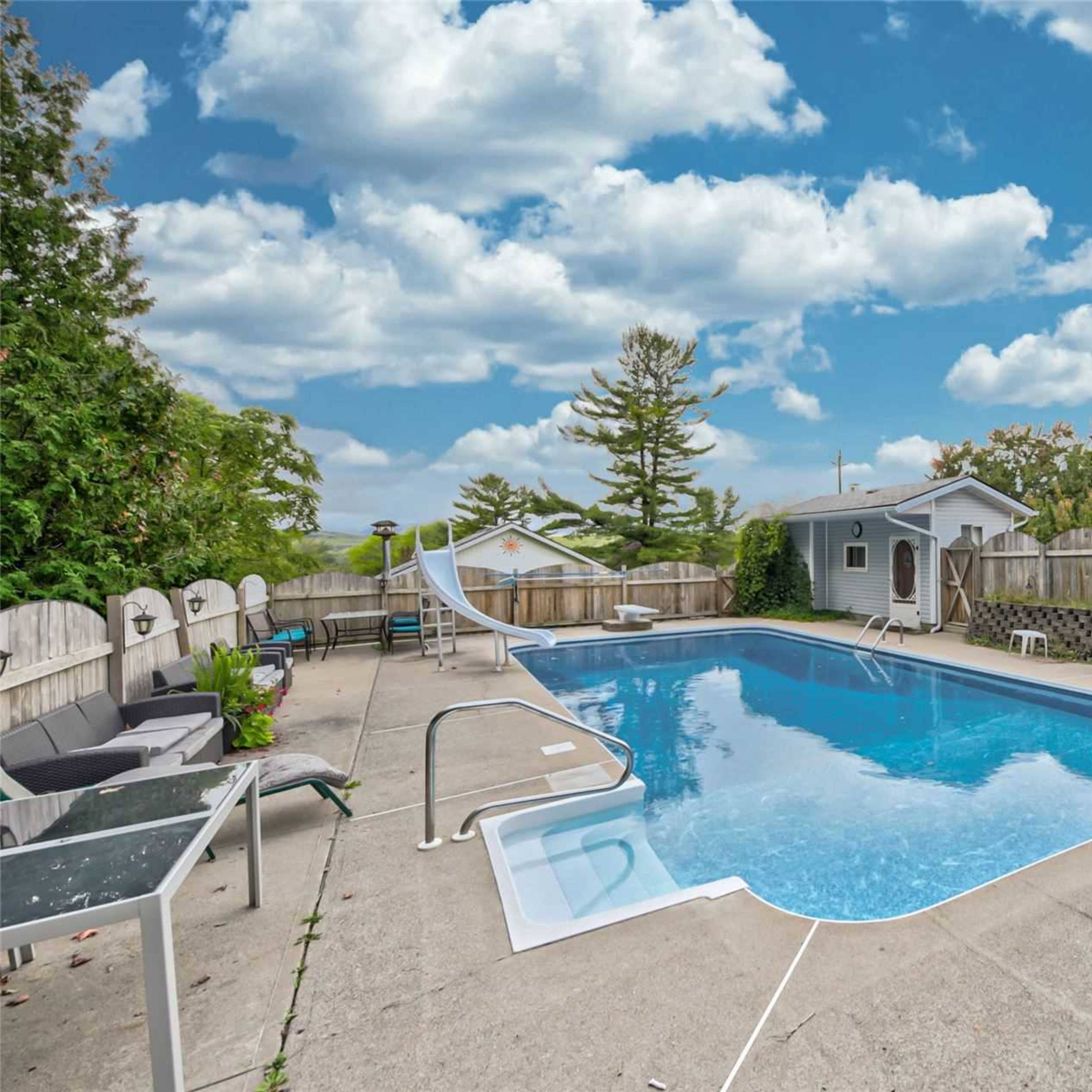 A swimming pool surrounded by lounge chairs and a slide, with a fence and trees in the background.