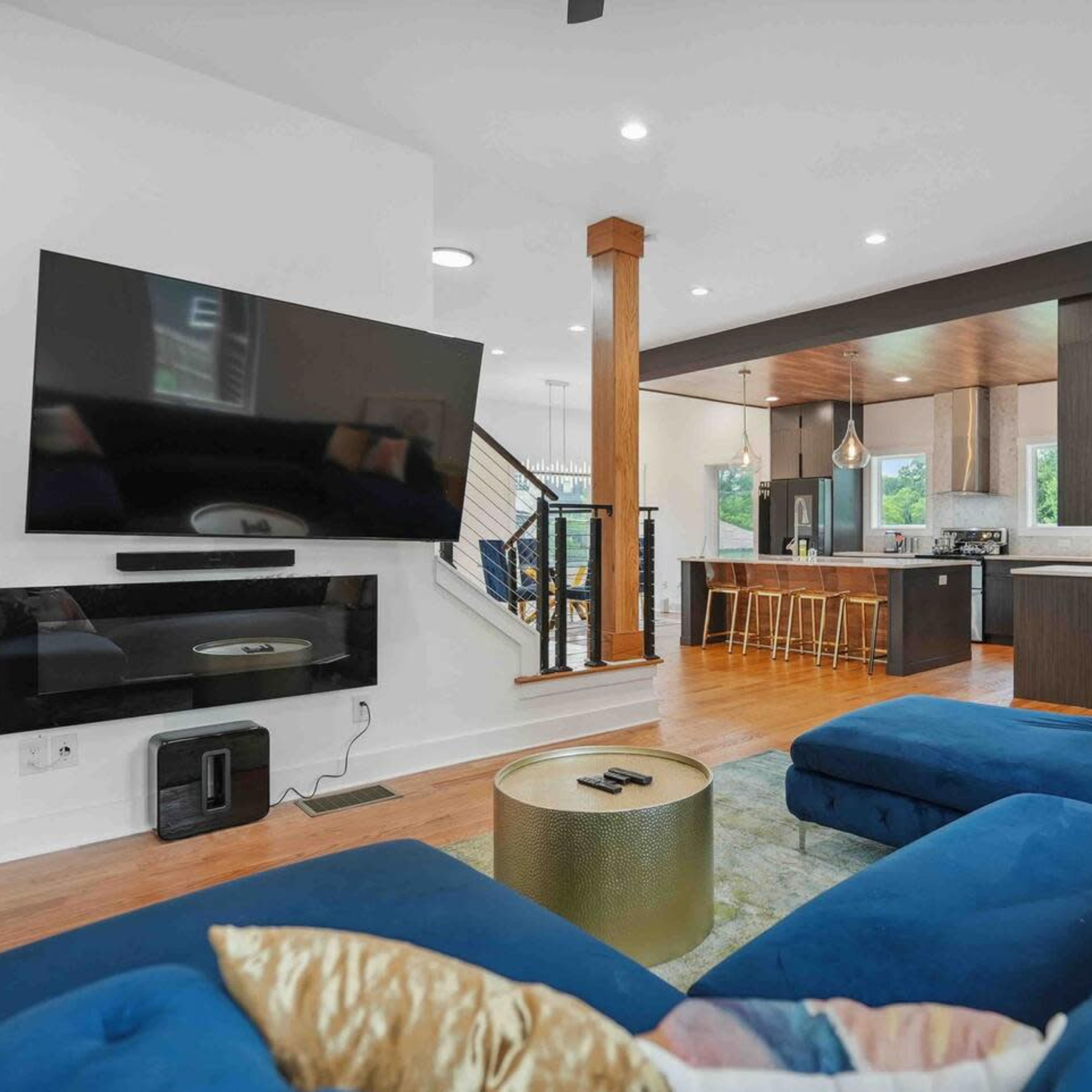 A modern living room featuring a blue sectional sofa, a large flat-screen TV mounted on the wall, and a kitchen area visible in the background with wooden bar stools.