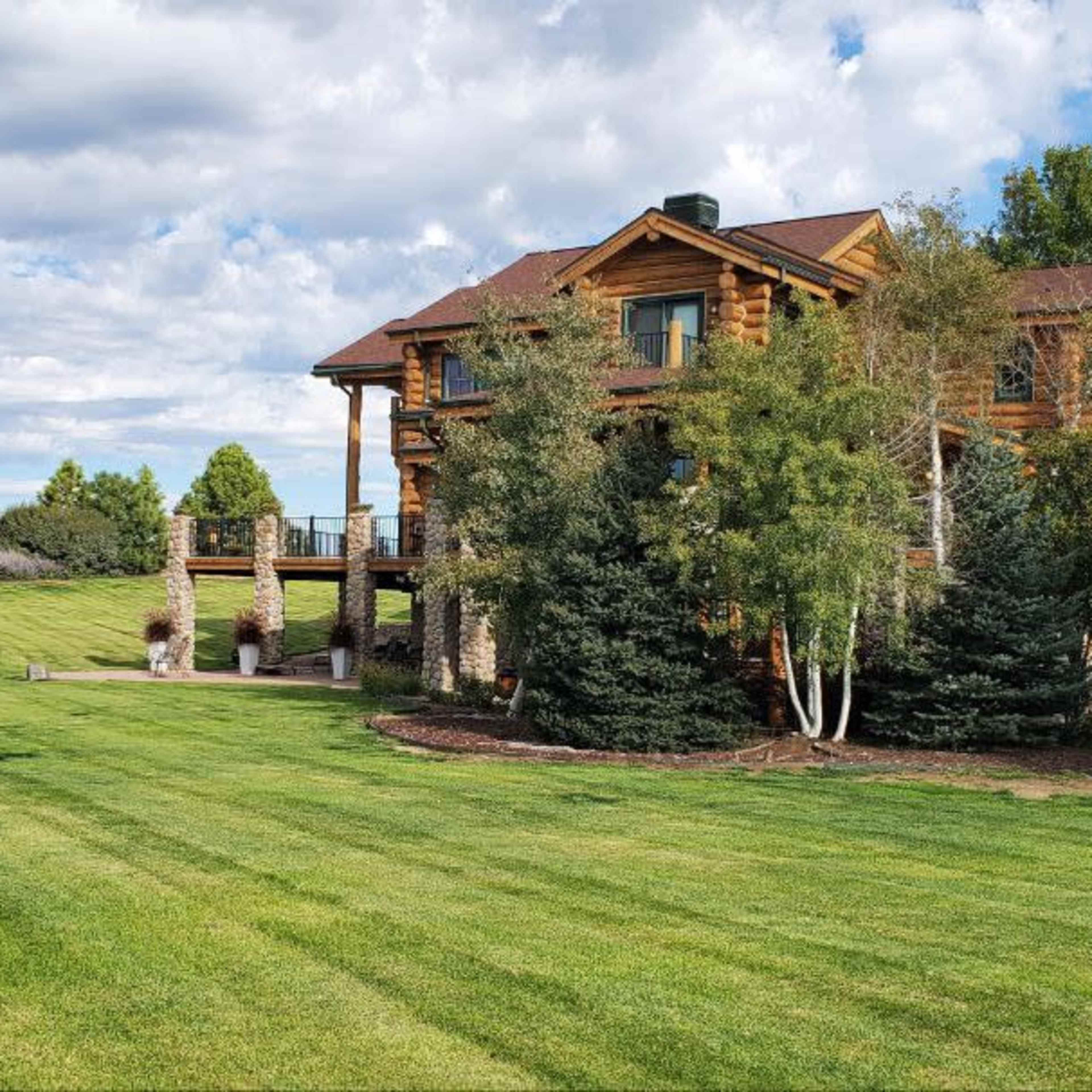 A large log cabin with a deck overlooks a manicured green lawn and landscaped garden under a partly cloudy sky.