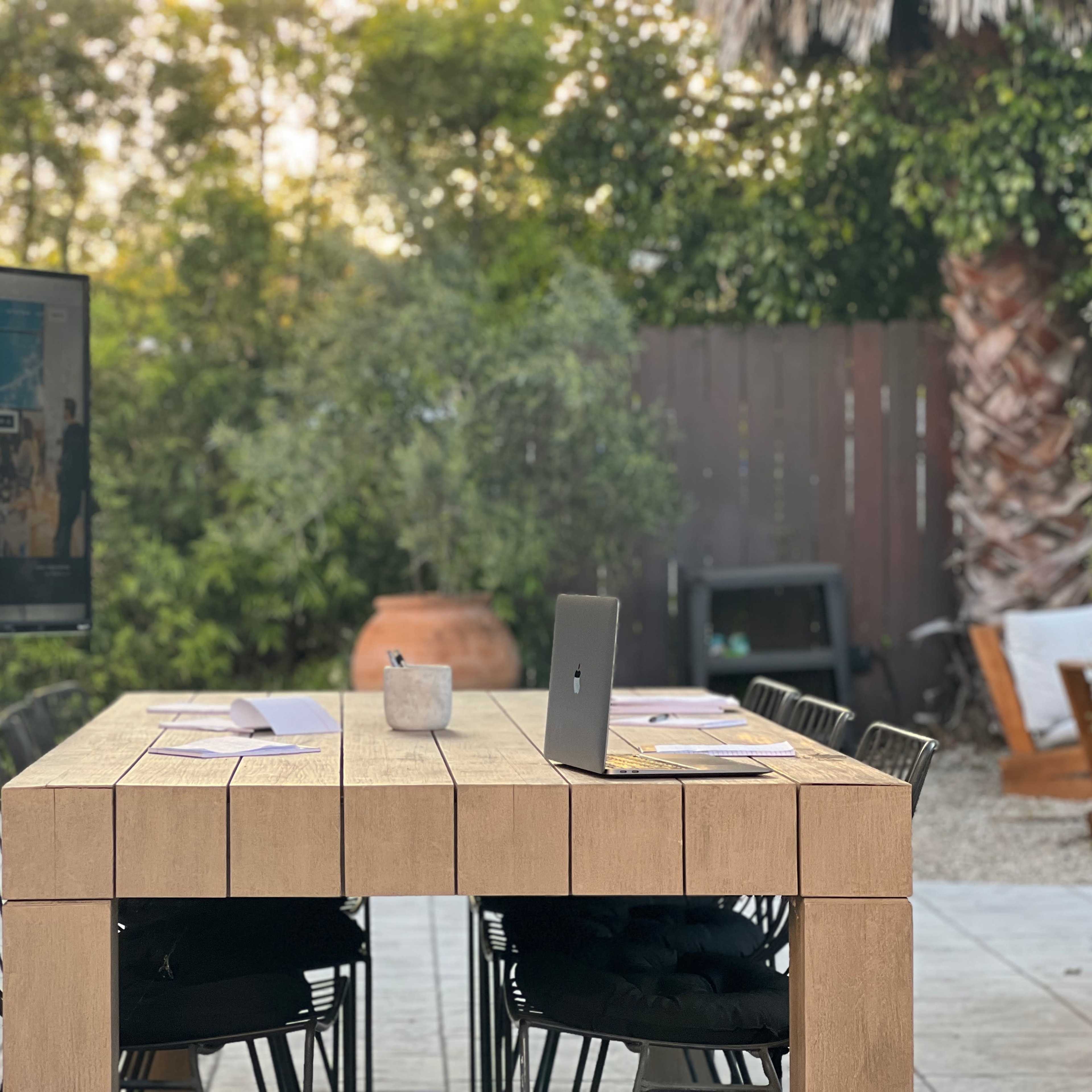 A large wooden table is set outdoors with chairs, accompanied by a laptop and a TV displaying a message about finding a space to fulfill a vision.