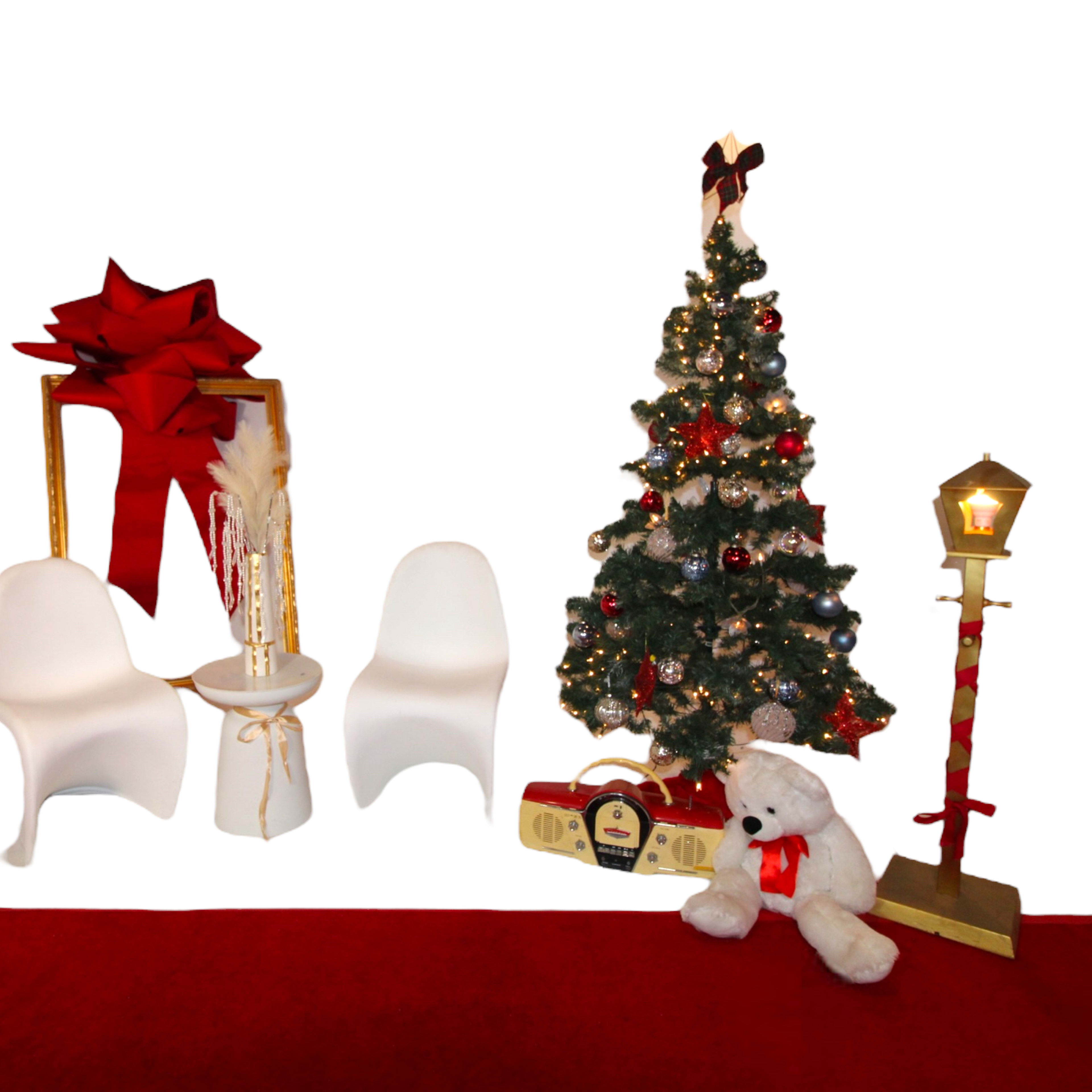 A festive arrangement with a decorated Christmas tree, two white chairs, a red backdrop, and various holiday-themed decorations, including a large bow and a teddy bear.