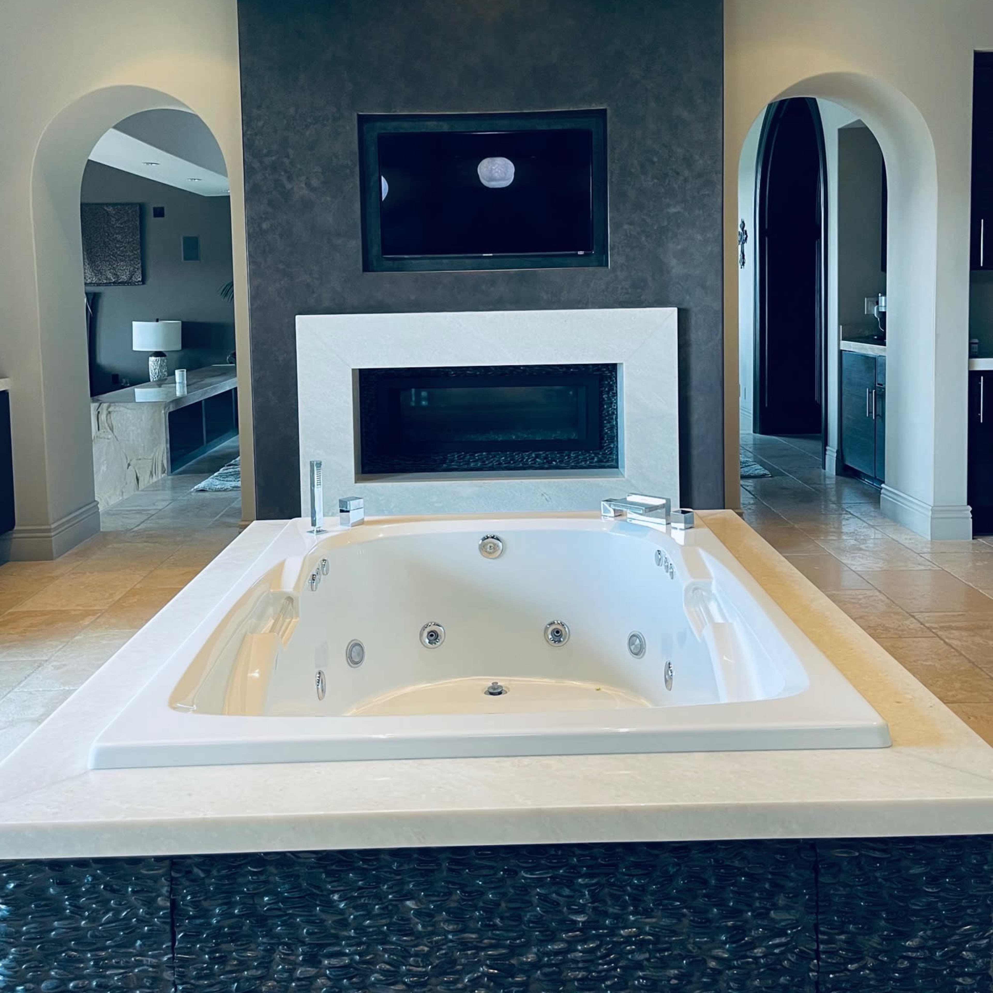 A modern bathroom featuring a large white jacuzzi tub, a flat-screen TV mounted on a wall, and a fireplace, all set against sleek gray and black finishes.