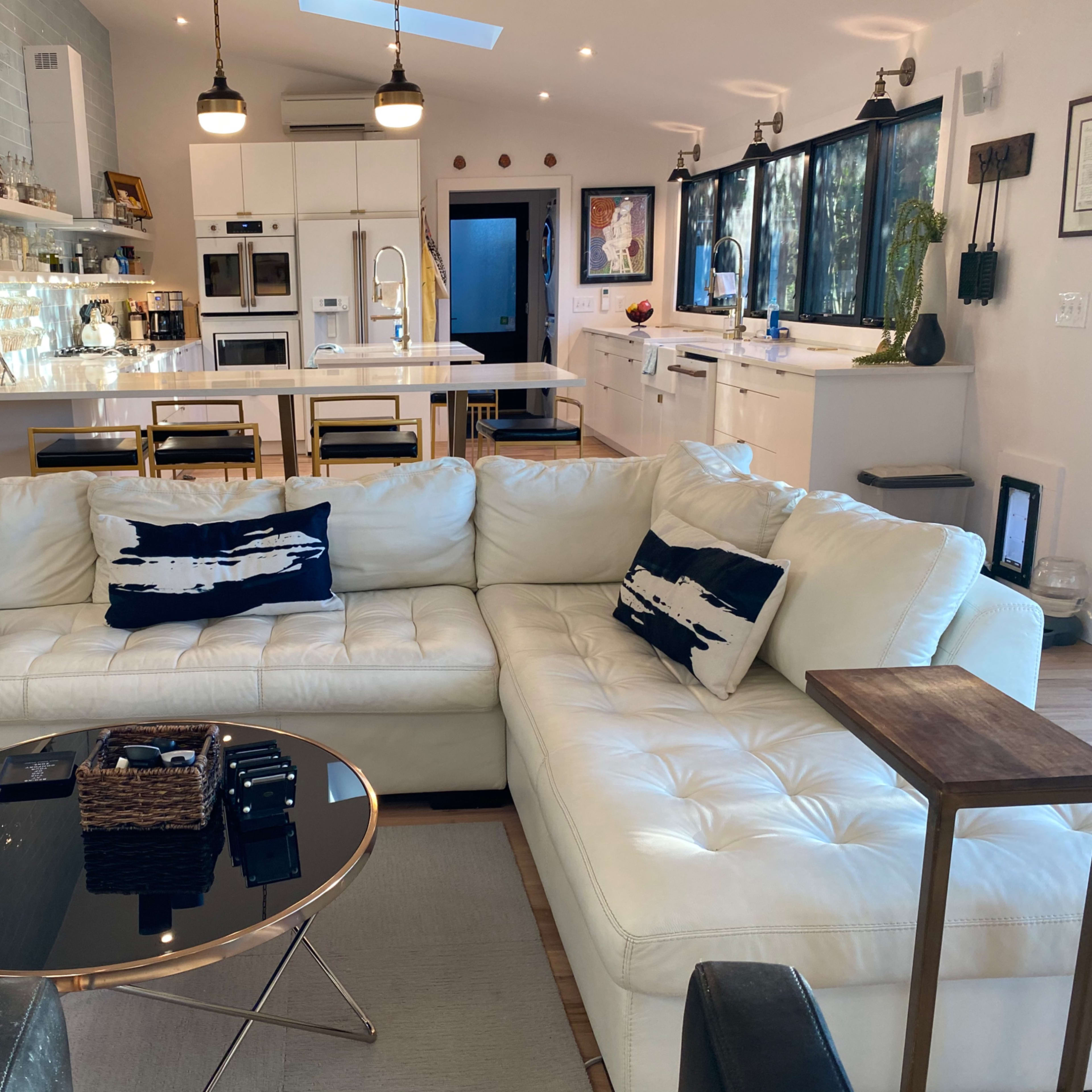 A modern, open-concept living area featuring a white sectional sofa, a black coffee table with a woven basket, and a kitchen in the background with white cabinetry and appliances.