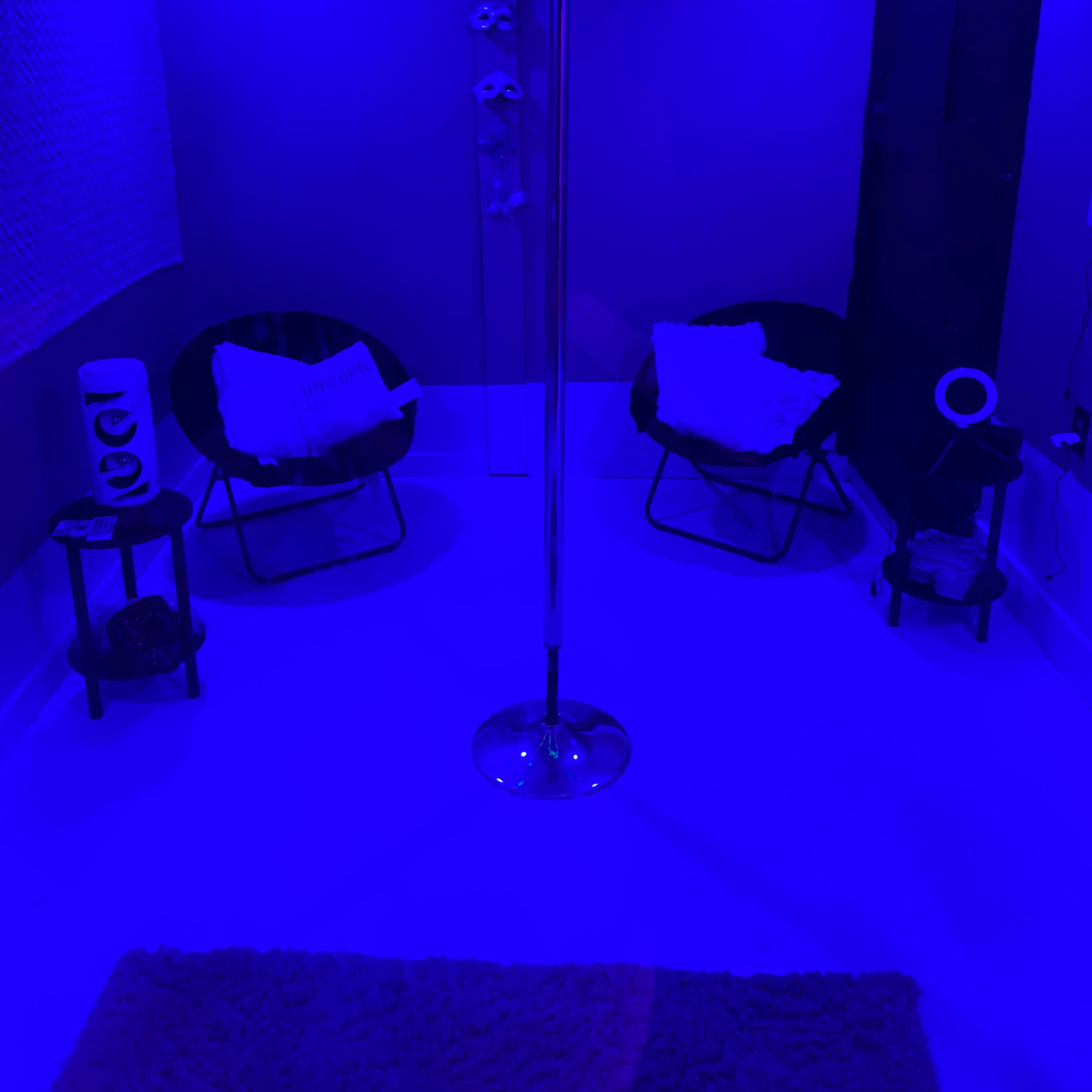 A small room features a dance pole in the center, surrounded by two black chairs, a small table, and soft lighting in shades of blue.