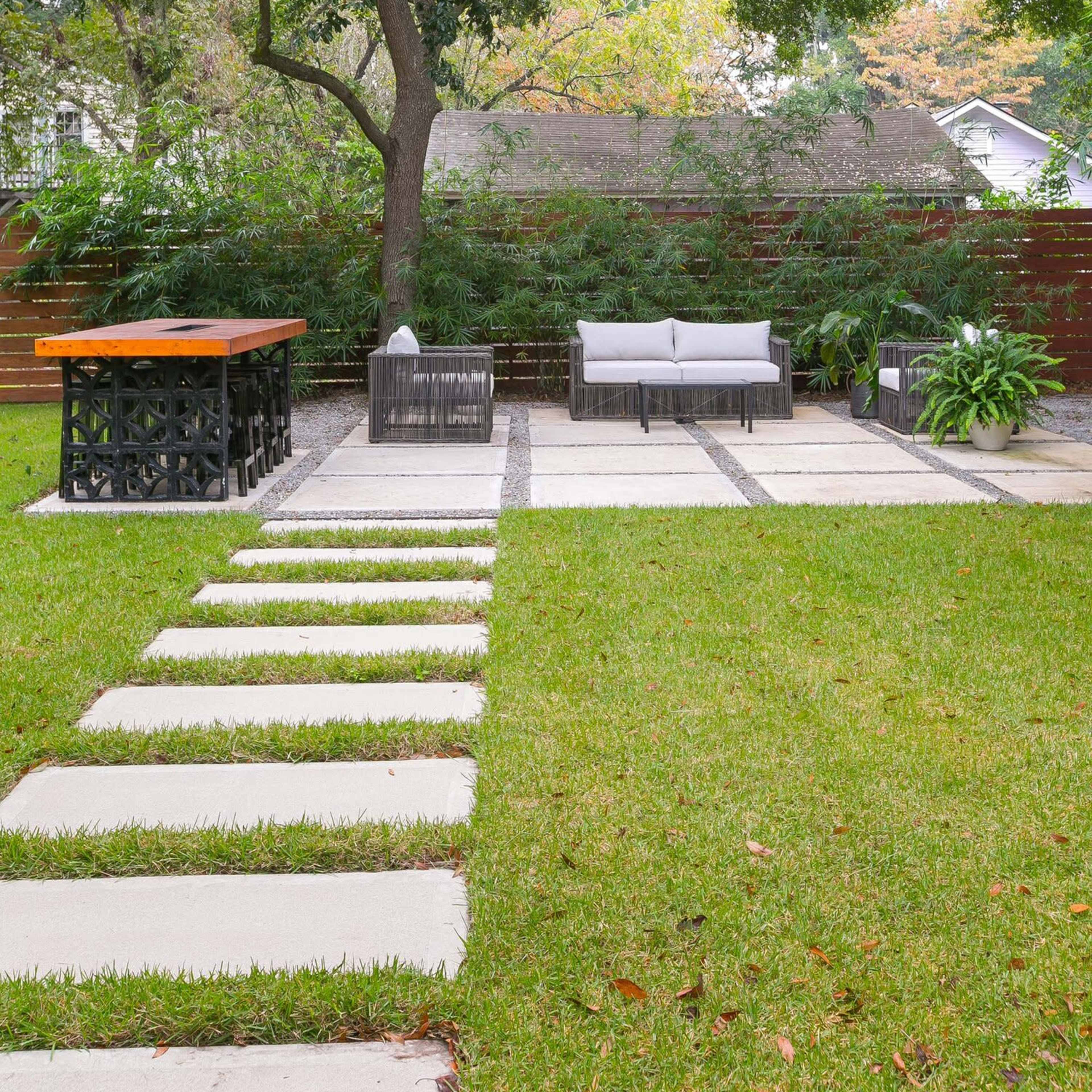 A backyard patio area featuring a stone pathway leading to a seating arrangement with a gray sofa and chairs, alongside a wood-topped bar table and landscaped greenery.