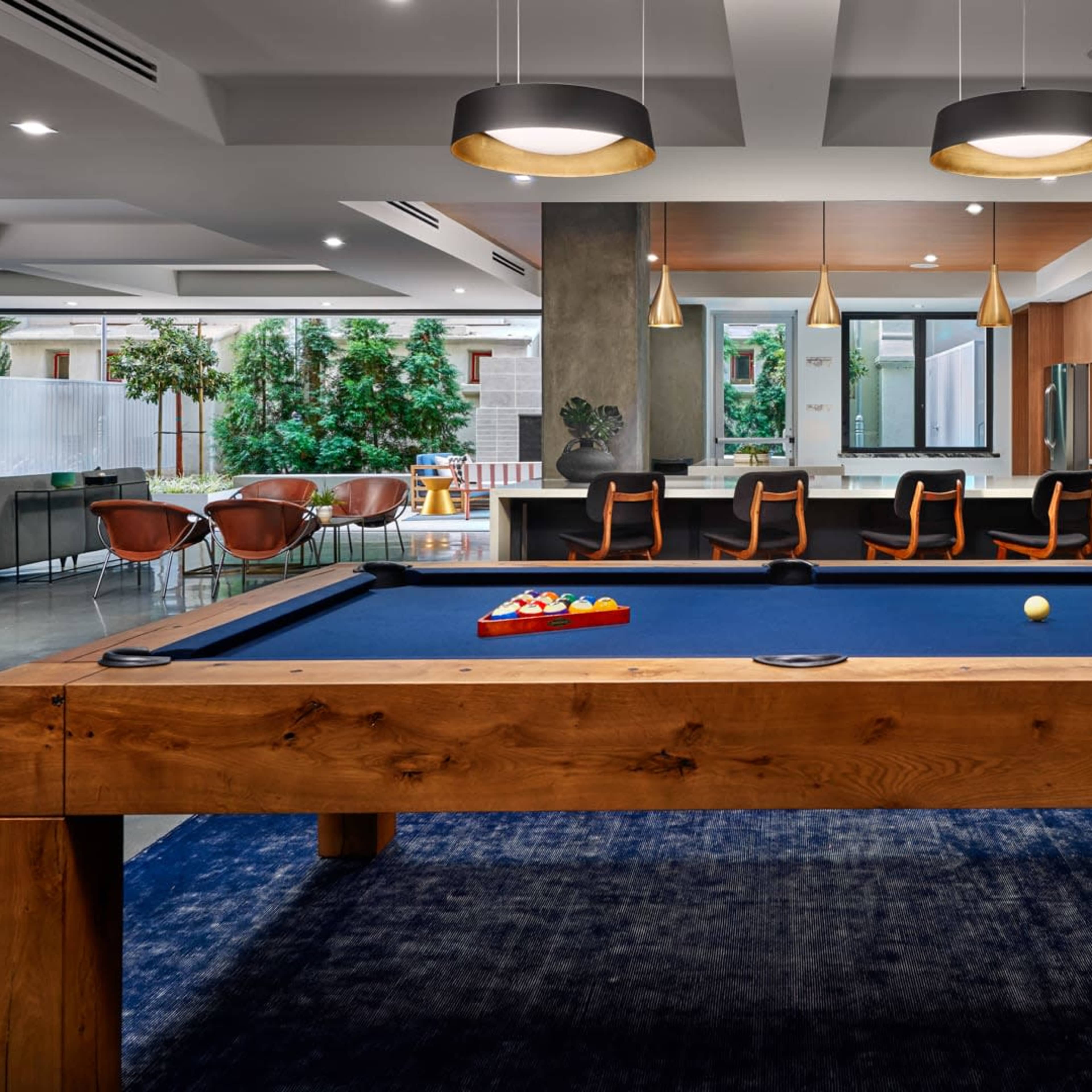 A modern game room featuring a large wooden pool table surrounded by a blue rug, with a kitchen and dining area visible in the background.