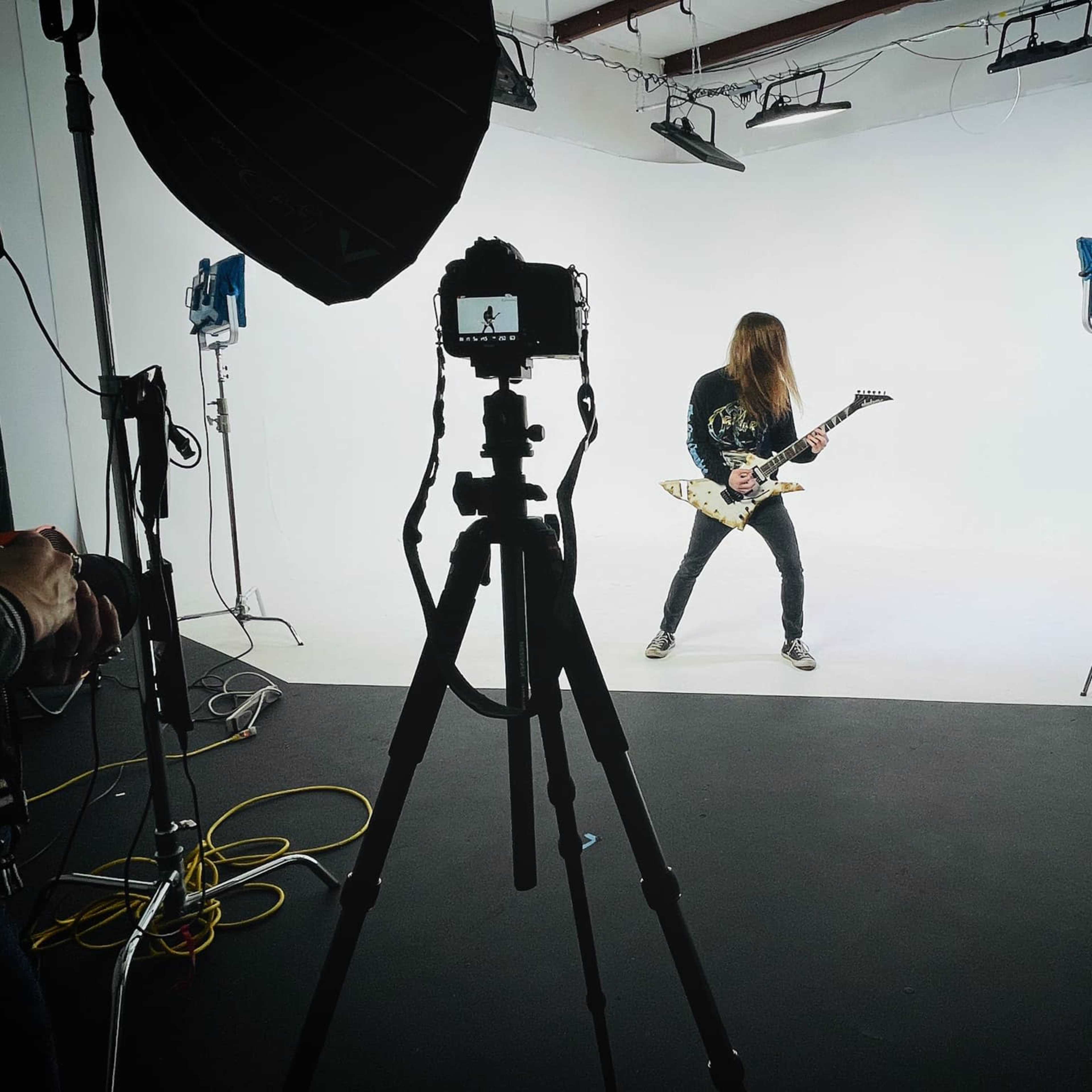 A guitarist stands in a studio space with a white backdrop, illuminated by several lights, while being photographed from a distance.
